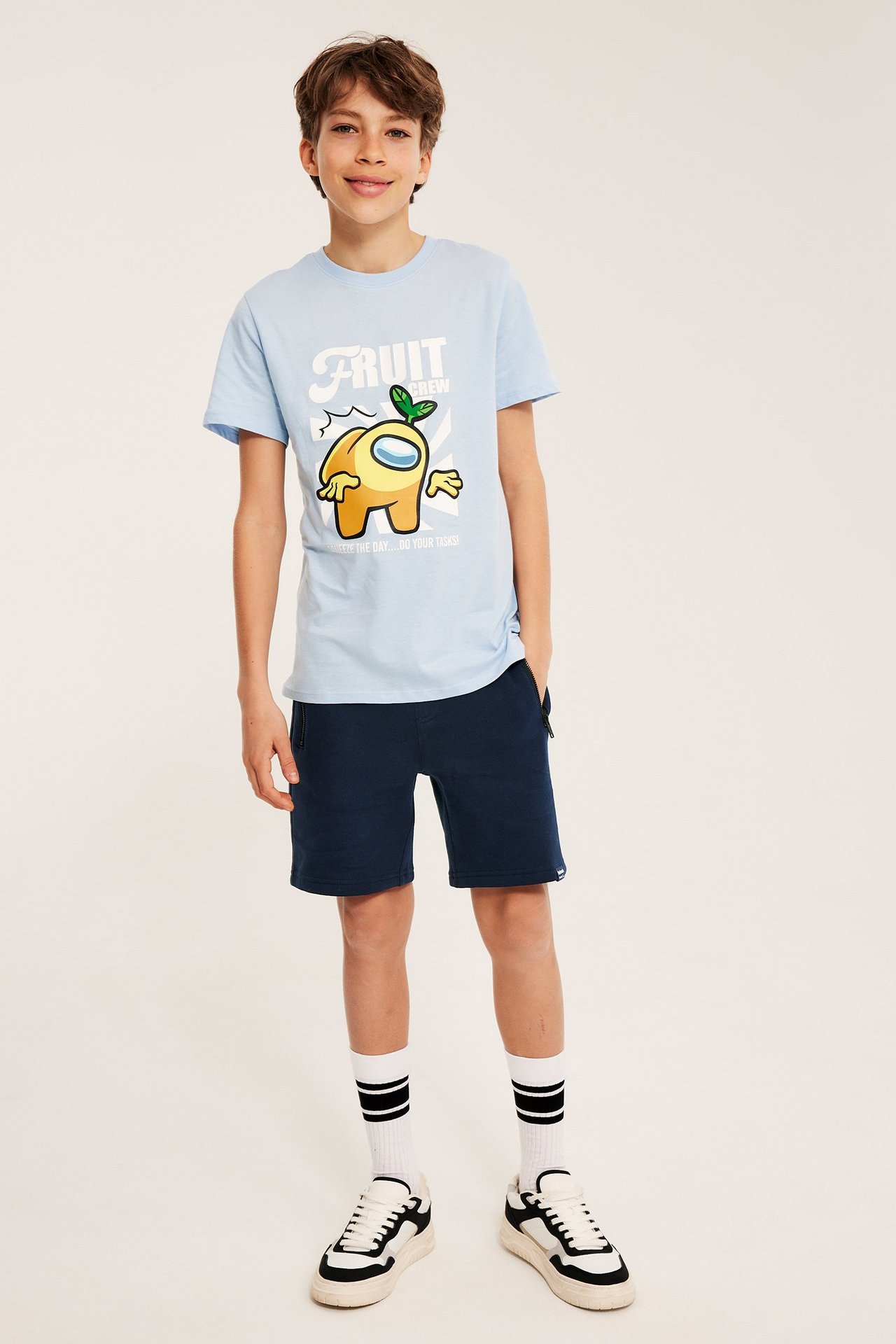 Widok z przodu: Dziecko w granatowych szortach dresowych, jasnoniebieskiej koszulce z nadrukiem i trampkach.
