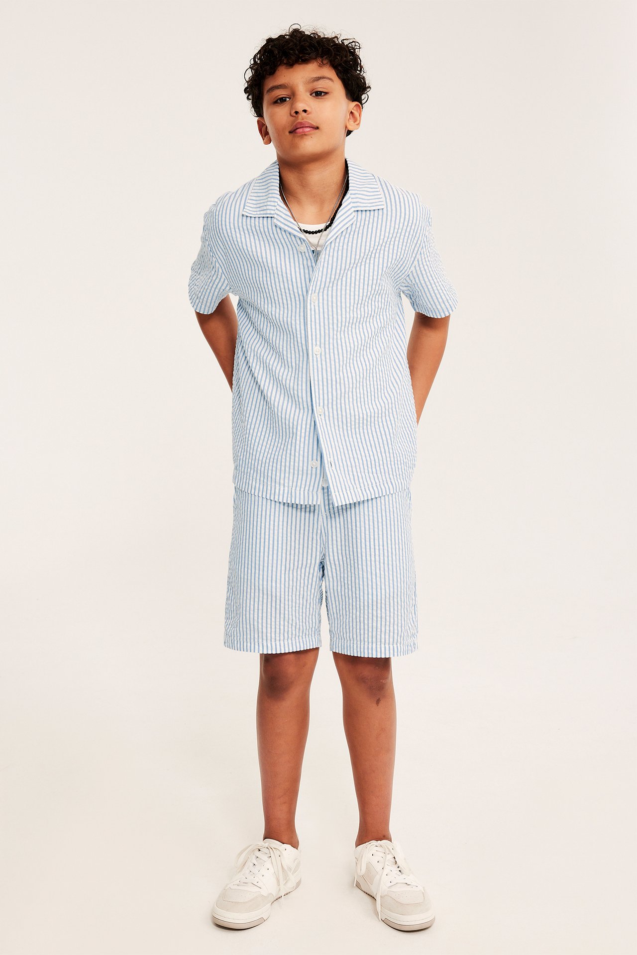 Dziecko od przodu: niebieska bawełniana koszula w paski z krótkim rękawem, pasujące szorty i tenisówki.