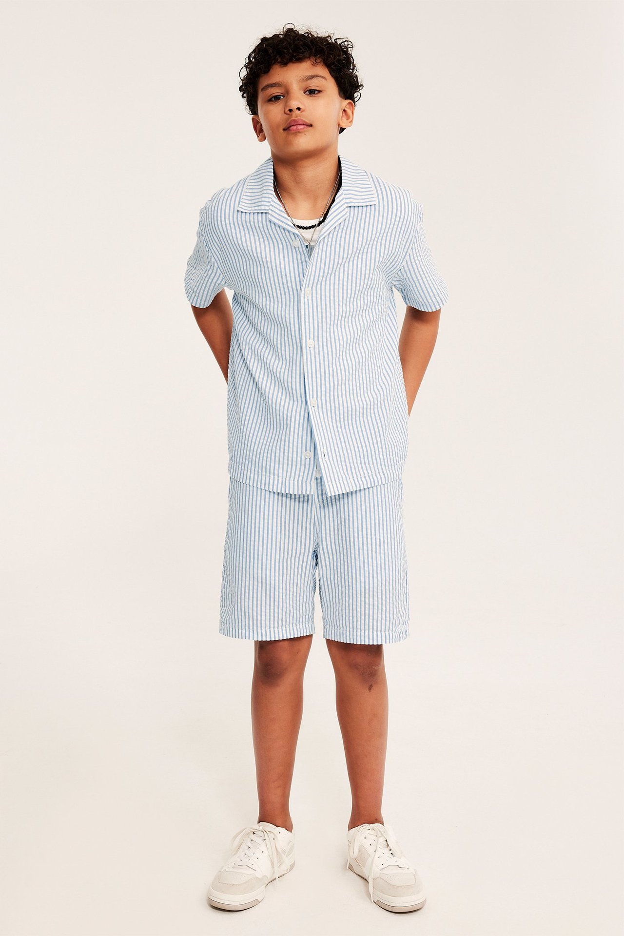 Dziecko od przodu: niebieska bawełniana koszula w paski z krótkim rękawem, pasujące szorty i tenisówki.