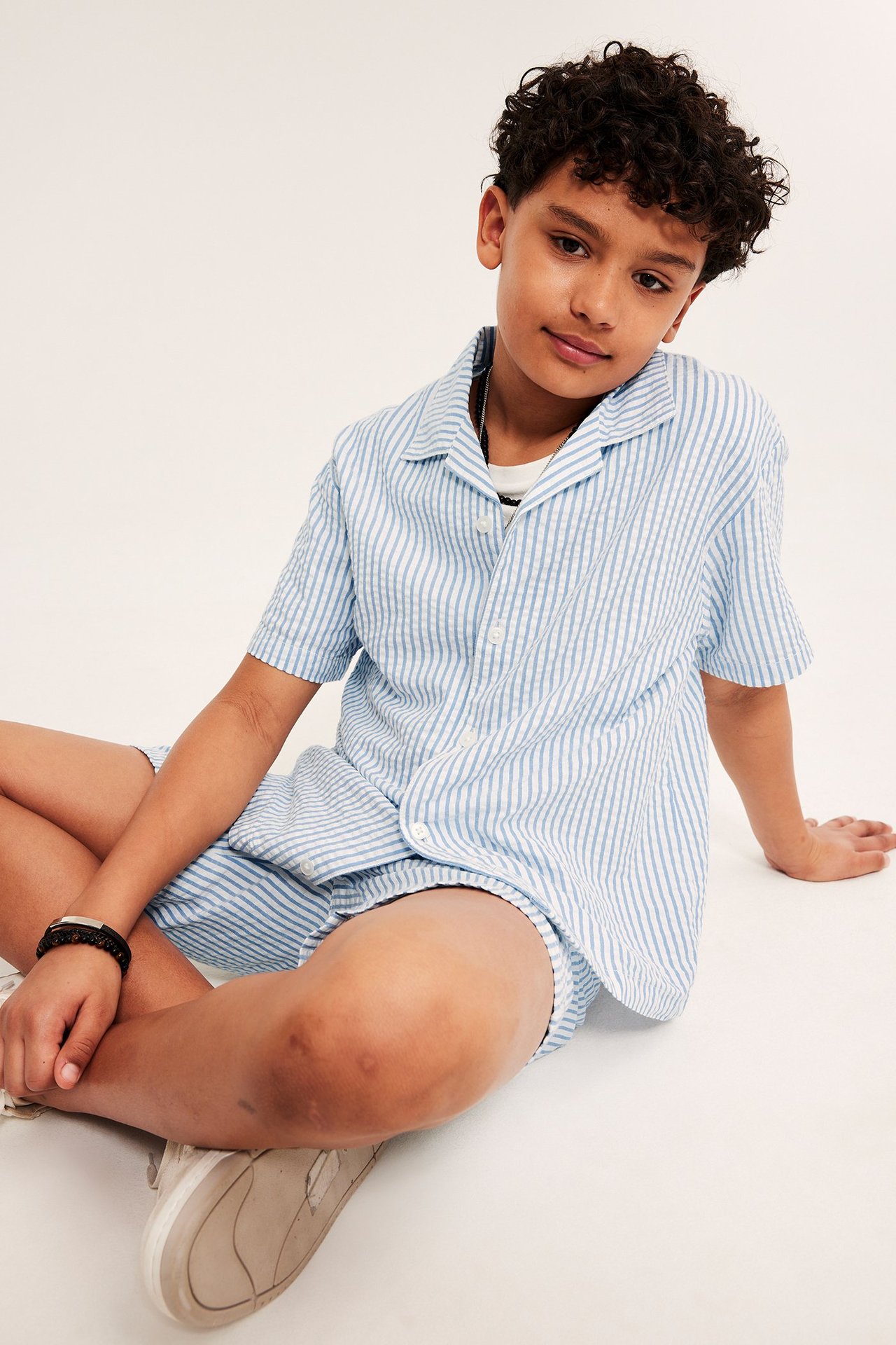 Widok z przodu: Dziecko w jasnoniebieskiej koszuli w paski seersucker, otwartej na białym t-shircie, z pasującymi szortami.
