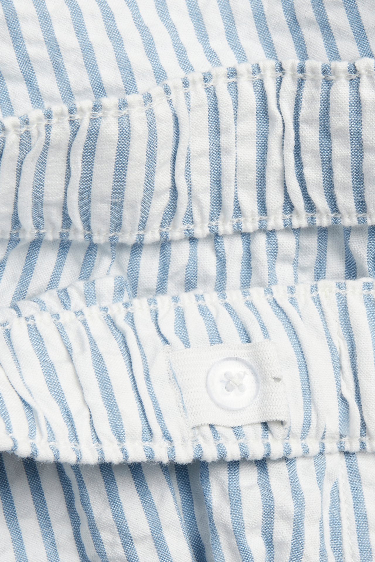 Zbliżenie na niebiesko-białe szorty z seersuckeru w paski, z elastycznym detalem i białym guzikiem.