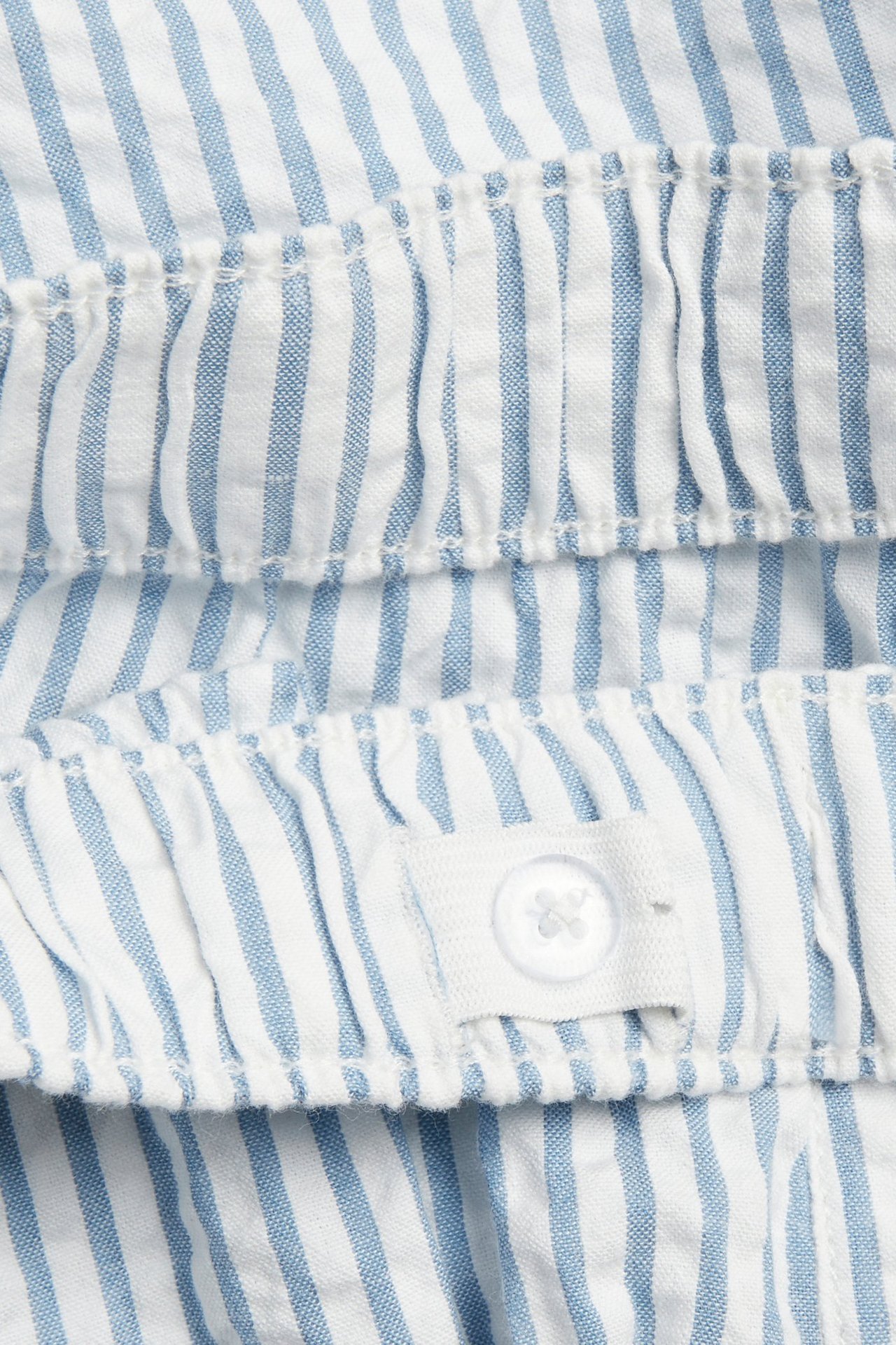 Zbliżenie na niebiesko-białe szorty z seersuckeru w paski, z elastycznym detalem i białym guzikiem.