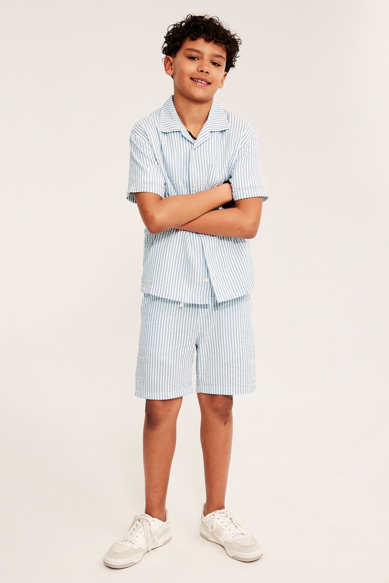 Widok z przodu dziecka: niebiesko-białe prążkowane szorty seersucker, pasująca koszula i białe sneakersy.