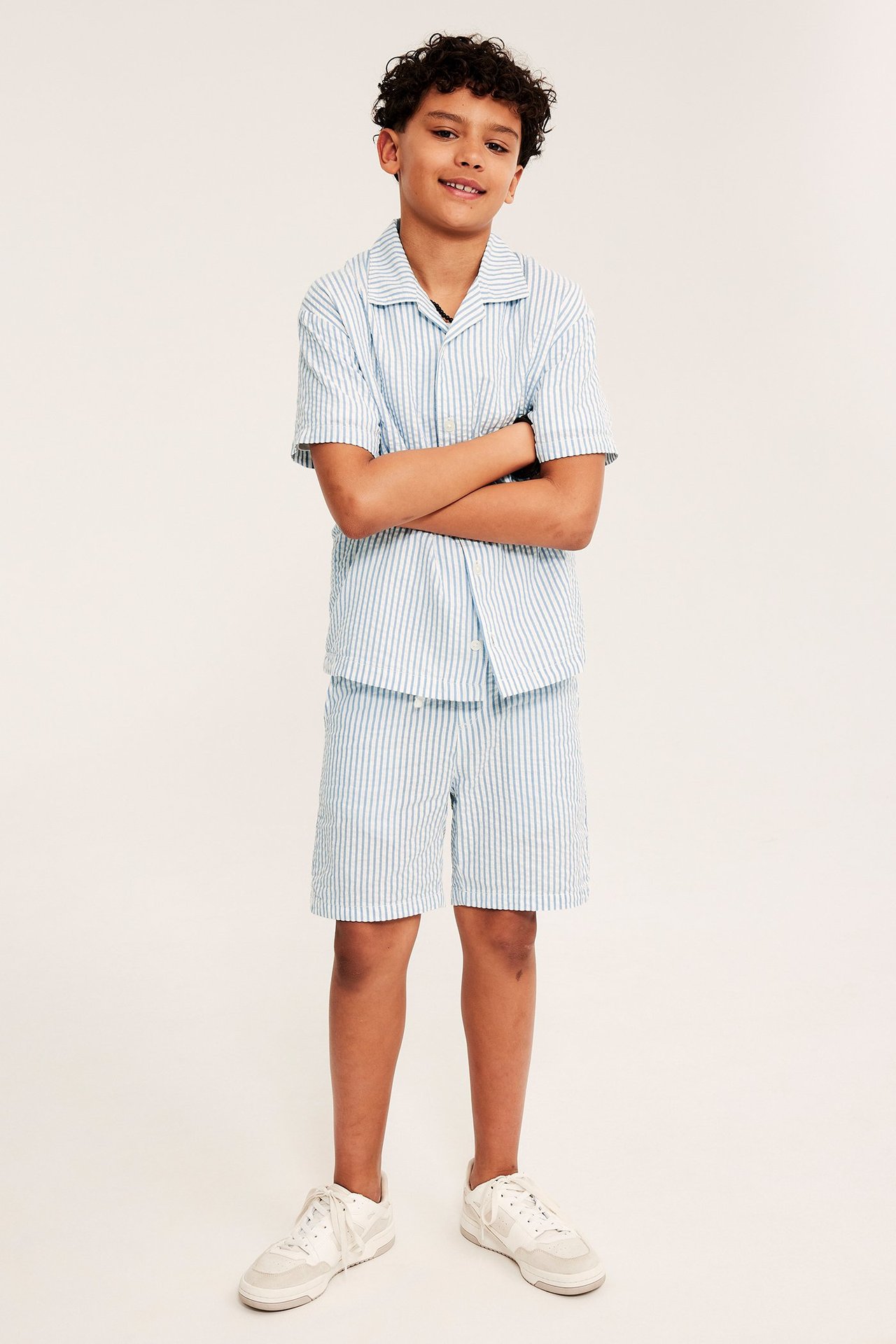 Widok z przodu dziecka: niebiesko-białe prążkowane szorty seersucker, pasująca koszula i białe sneakersy.