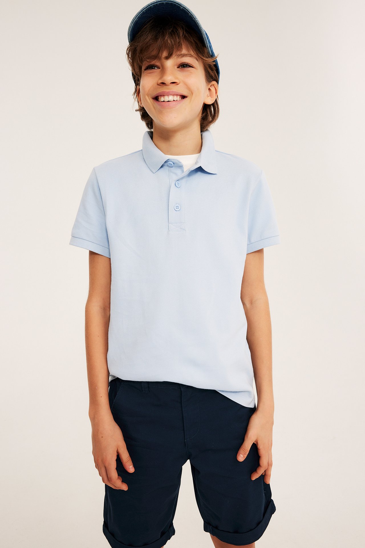 Lapsi edestä tummansinisissä chinoshortseissa, vaaleansinisessä pikeepaidassa ja tummansinisessä lippalakissa.
