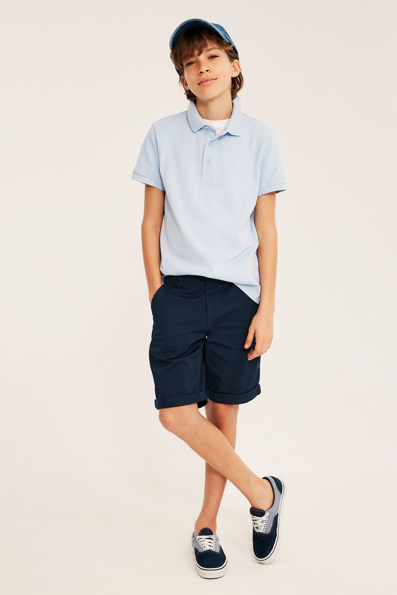 Edestä: Lapsi yllään tummansiniset puuvillachinoshortsit, vaaleansininen poolopaita, farkkulippis ja siniset tennarit.