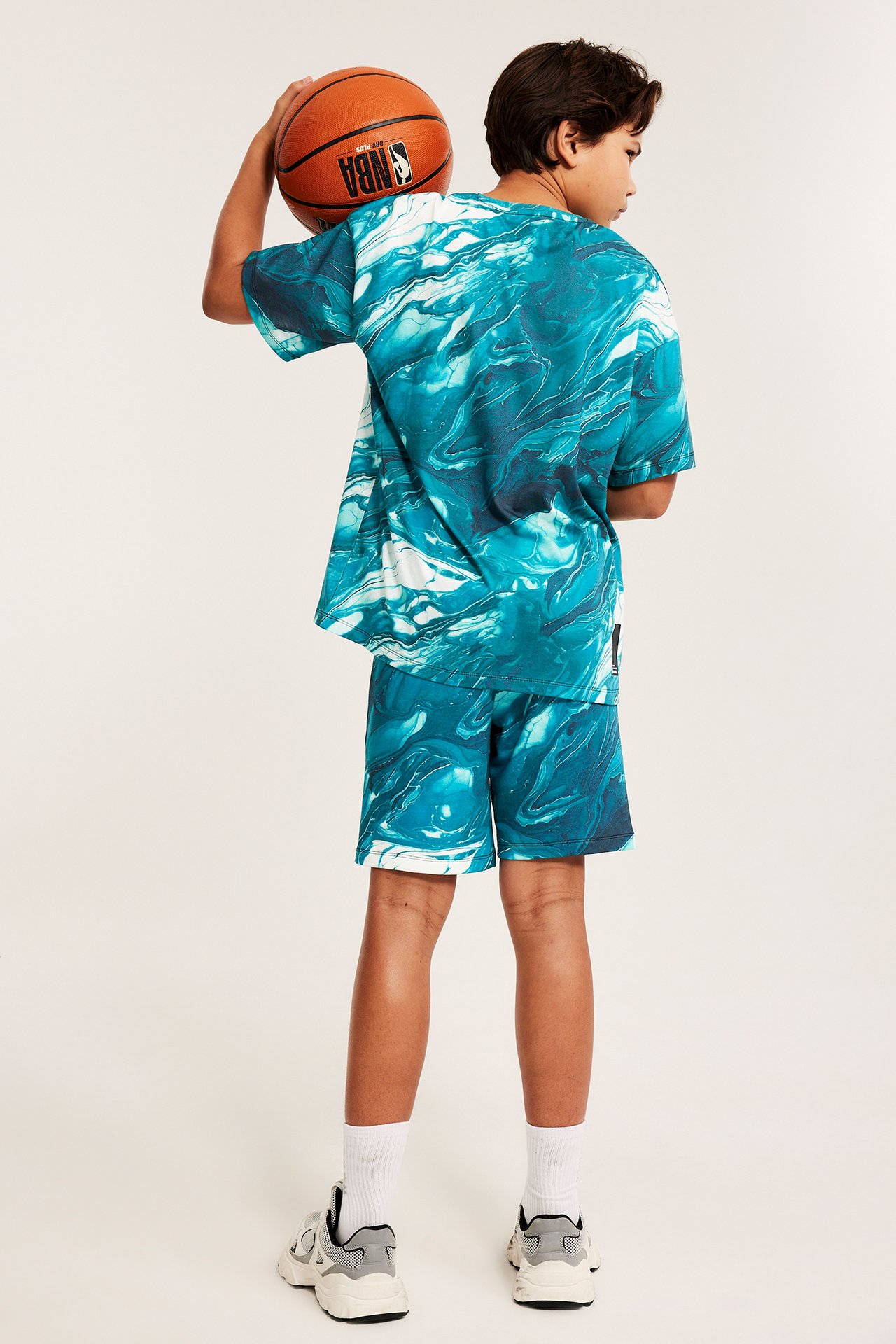 Lapsi takaa turkoosinvalkoisissa marmorikuvioisissa shortsissa ja paidassa, pitelemässä koripalloa.