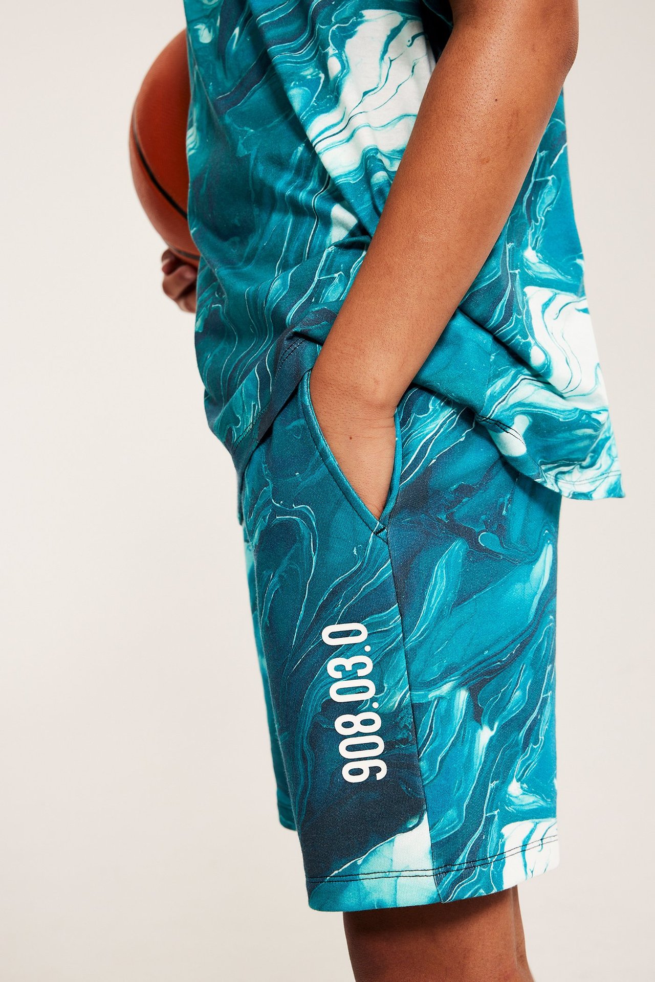 Sivukuva lapsesta, yllään petrolinsiniset ja luonnonvalkoiset kuviolliset college-shortsit valkoisilla numeroilla, t-paita, koripallo kädessä.