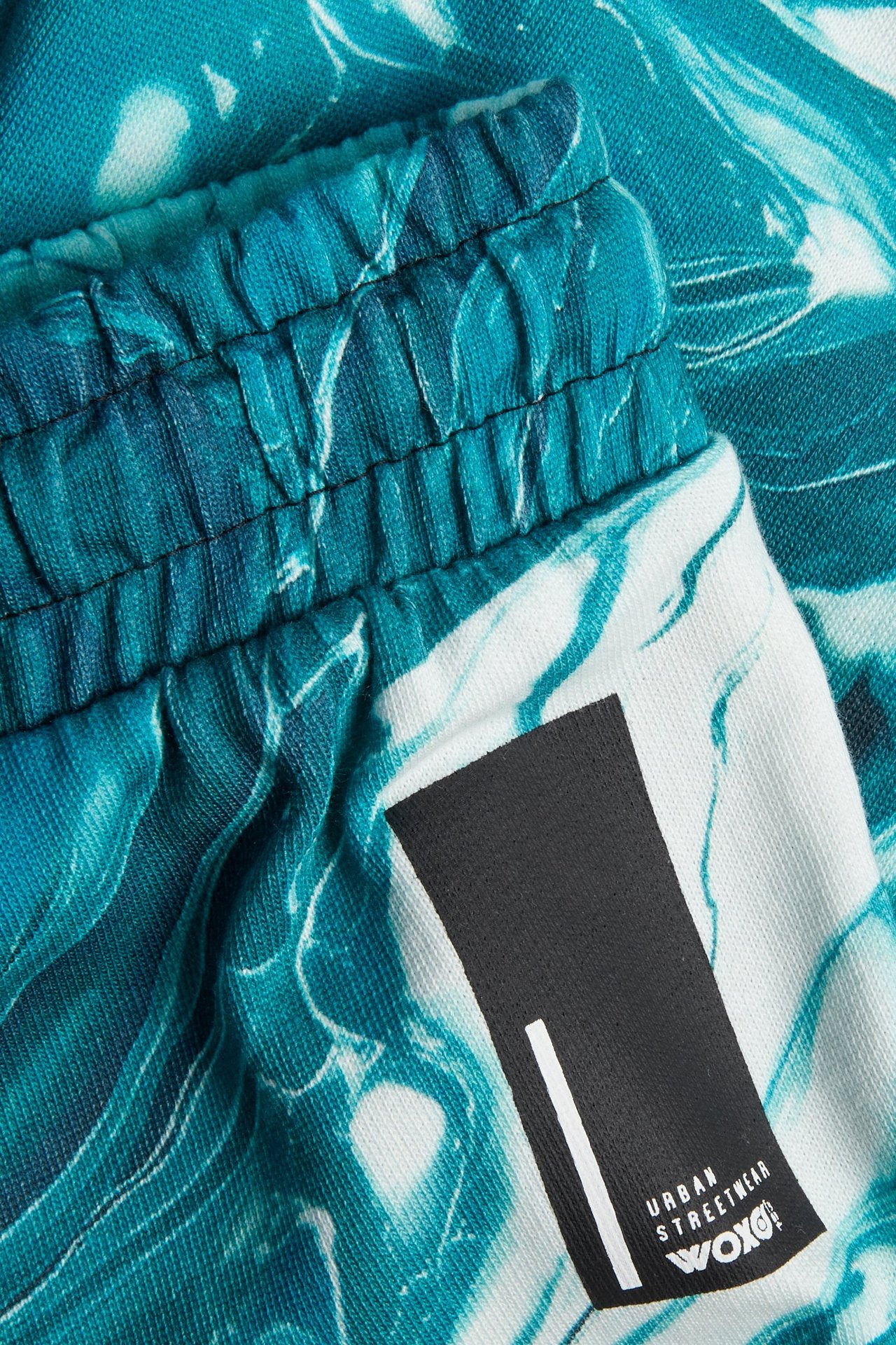 Zbliżenie: Bawełniane spodenki dresowe w kolorze ecru, z elastyczną talią i turkusowym, abstrakcyjnym wzorem.