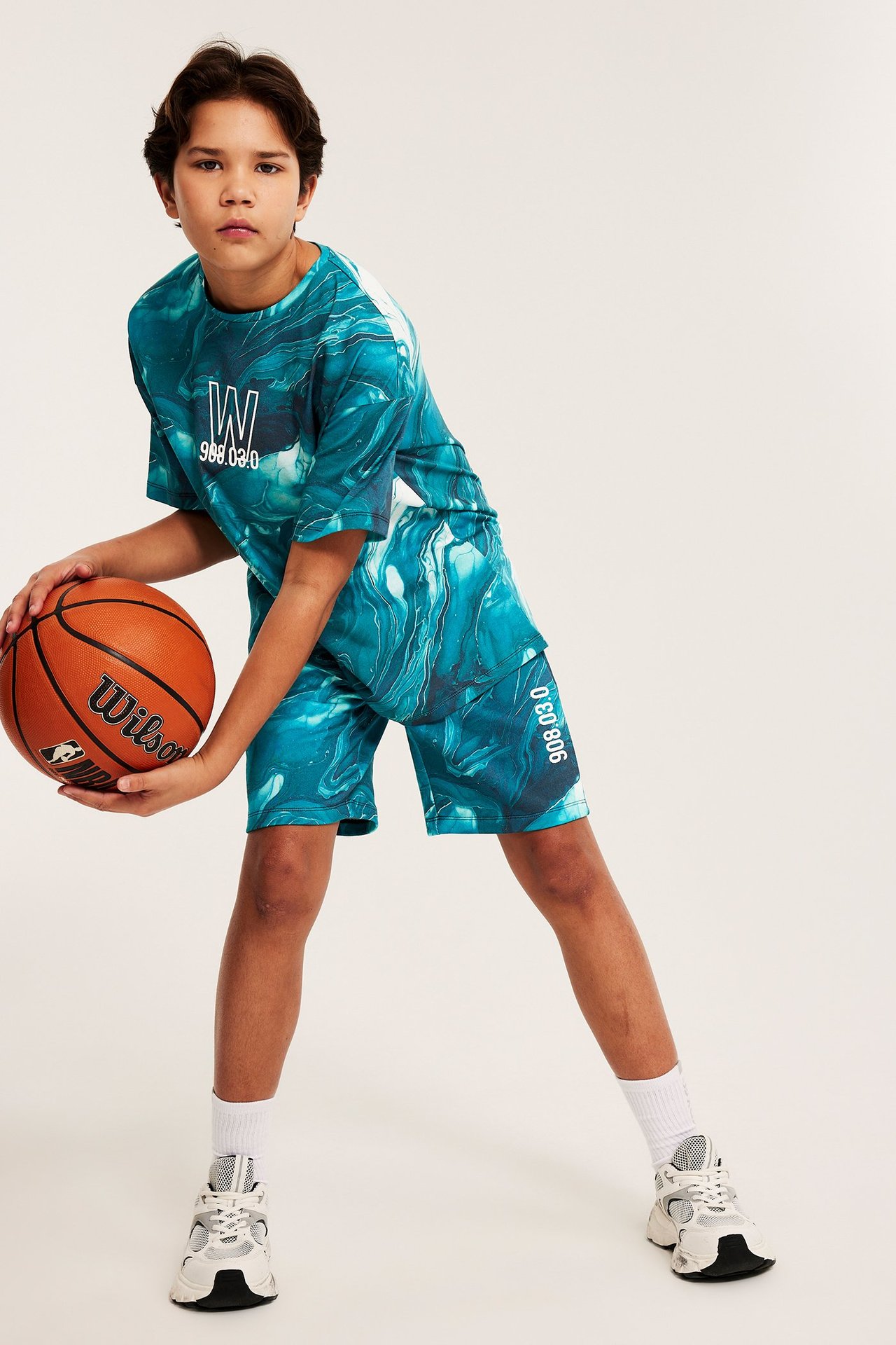 Widok z przodu: dziecko w turkusowo-białych wzorzystych szortach, t-shircie i sneakersach, trzymające piłkę.