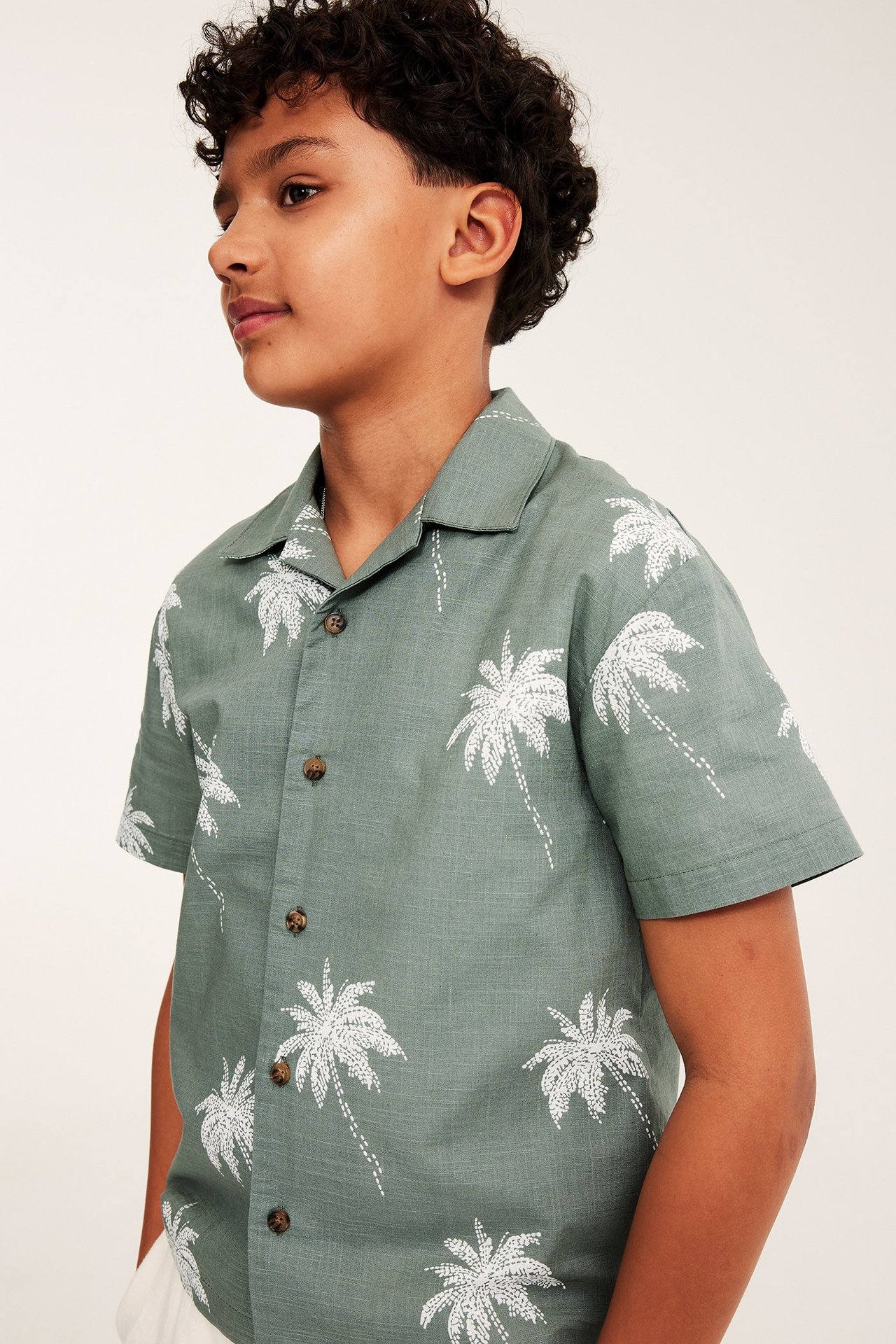 Sidovy av ett barn i en grön resortskjorta i bomull med vita palmer och bruna knappar.