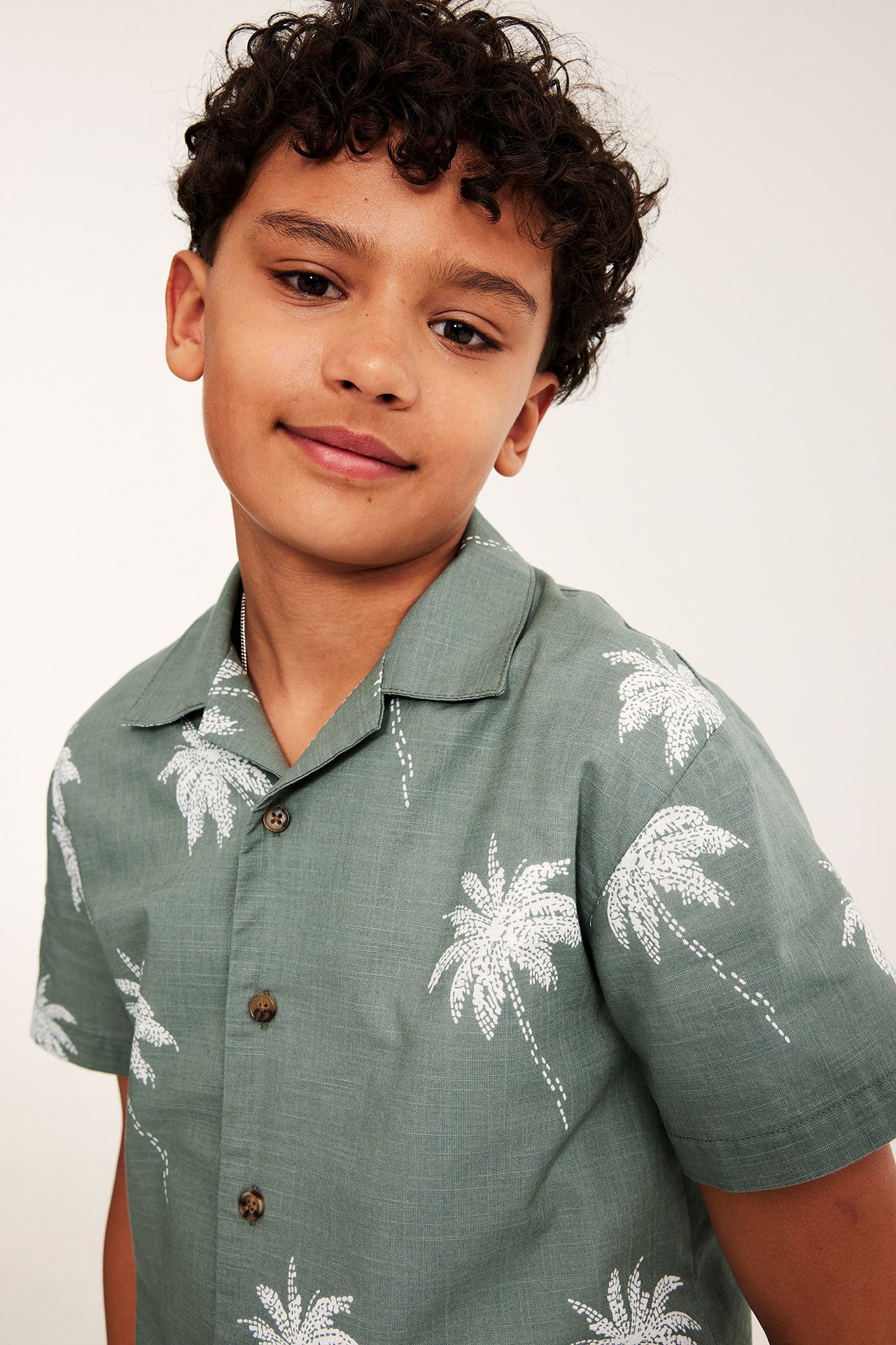 Närbild, framifrån, av en kille i en grön bomullsskjorta med vitt palmmönster och bruna knappar.