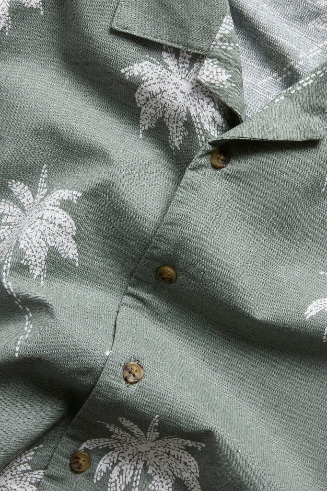 Zbliżenie na zieloną bawełnianą koszulę resortową z białym nadrukiem palm i brązowymi guzikami.