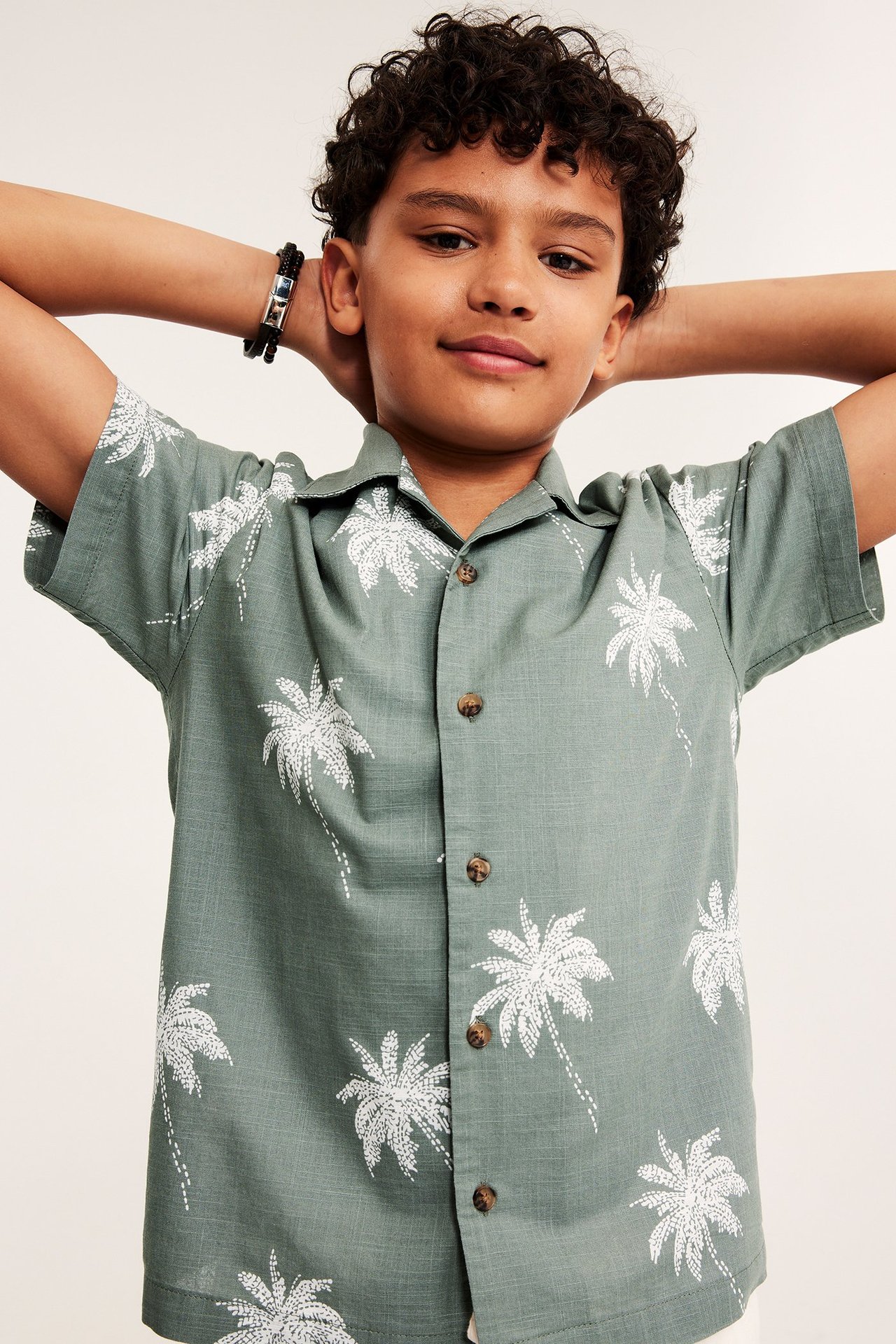 Frontvy: Pojke i grön bomullsskjorta med vitt palmtryck, bruna knappar och ett svart armband.