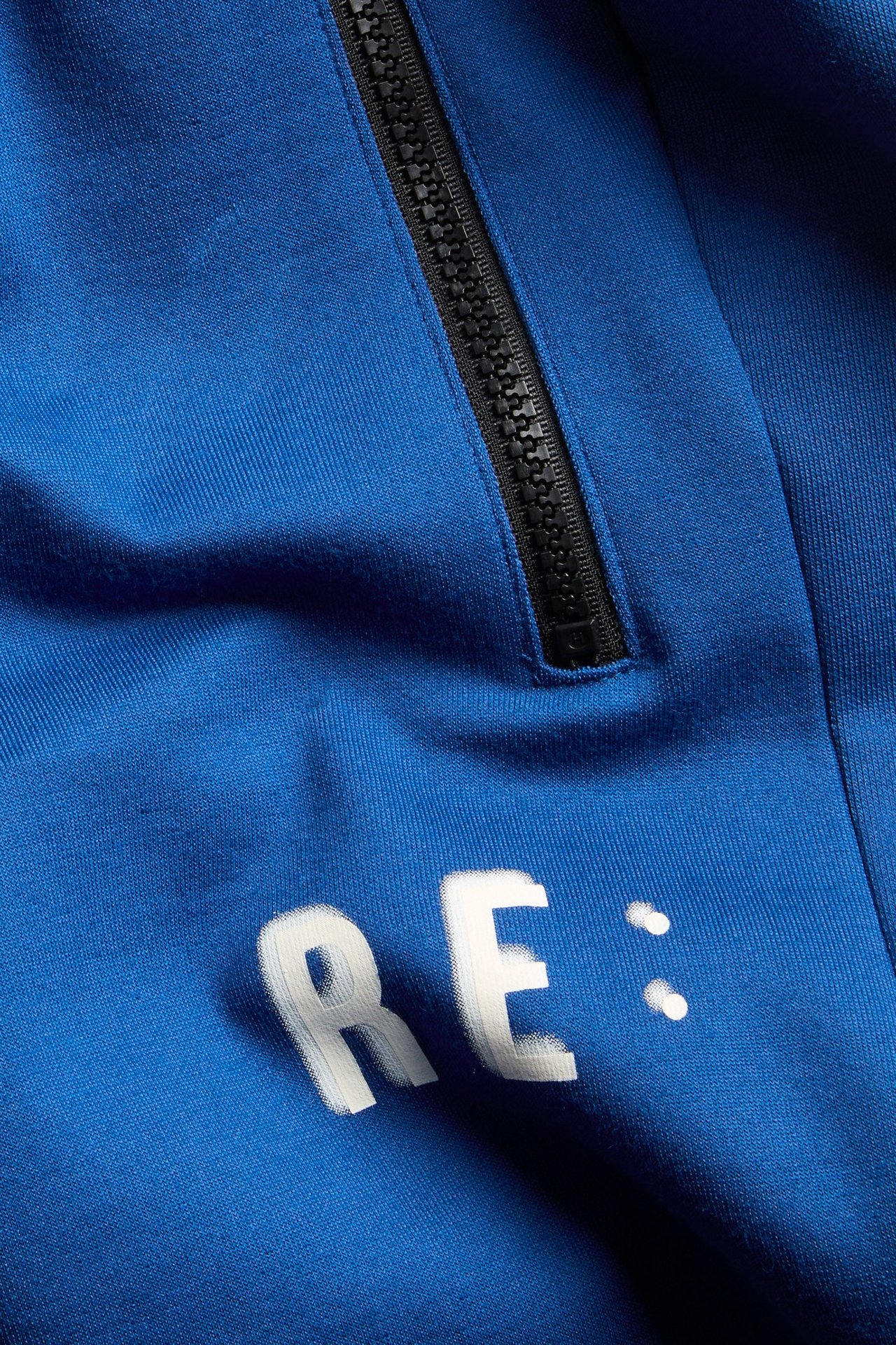 Närbild av mörkblå interlockshorts med synlig svart dragkedja och vitt "RE:" textprint.
