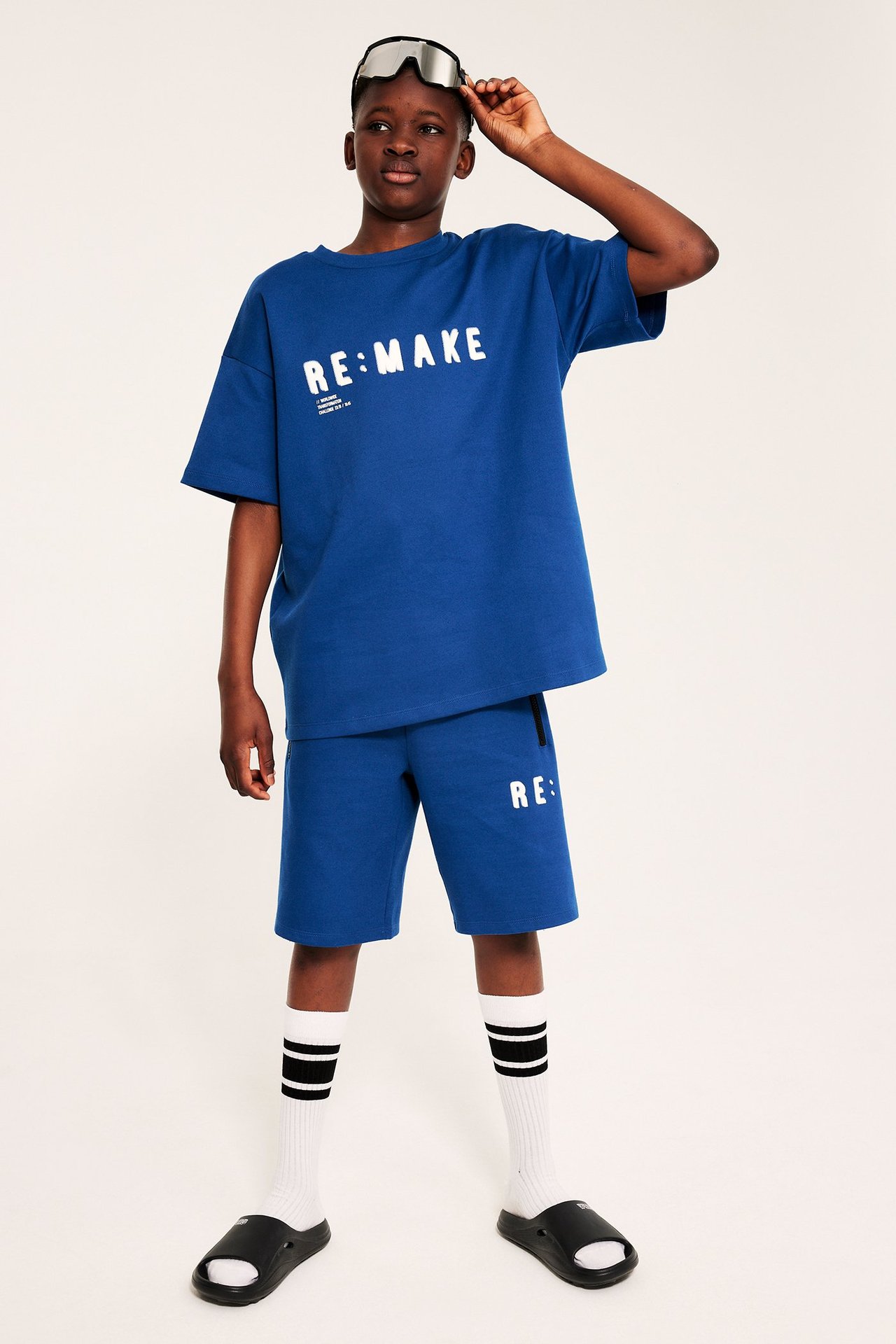 Dziecko nosi ciemnoniebieskie szorty z nadrukiem, pasujący T-shirt, białe skarpetki, czarne klapki i gogle.
