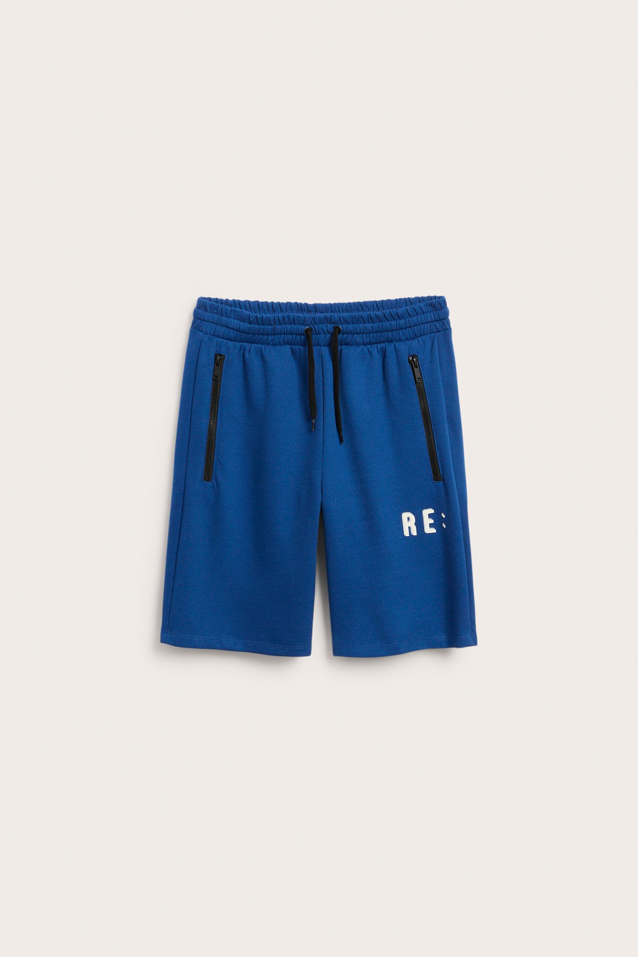 Framifrån, blå sportiga shorts med dragsko, svarta dragkedjefickor och ett vitt 'RE:'-tryck.