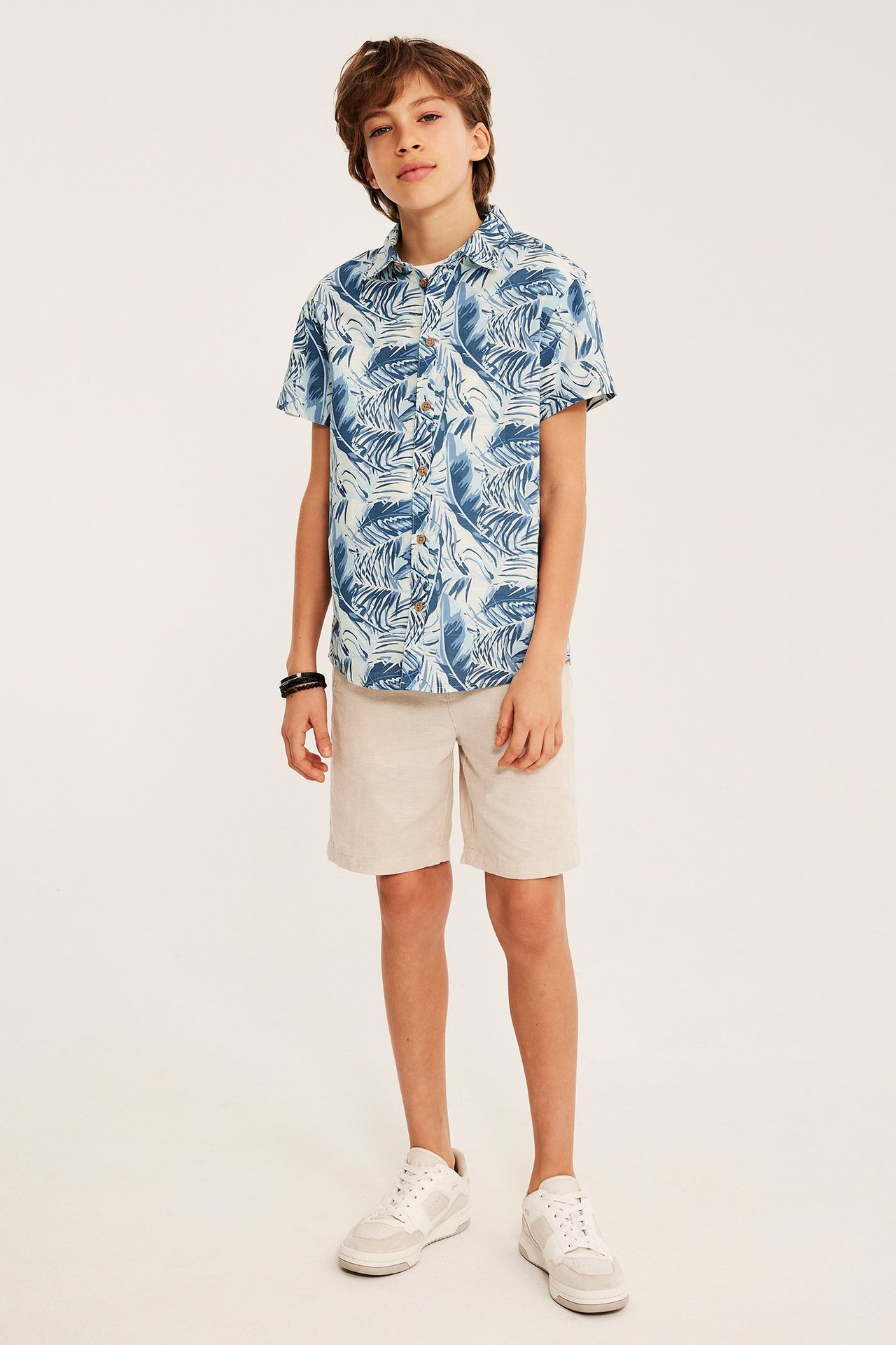 Framifrån: Ett barn i blå- och offwhite-mönstrad kortärmad skjorta, beiga shorts och vita sneakers.