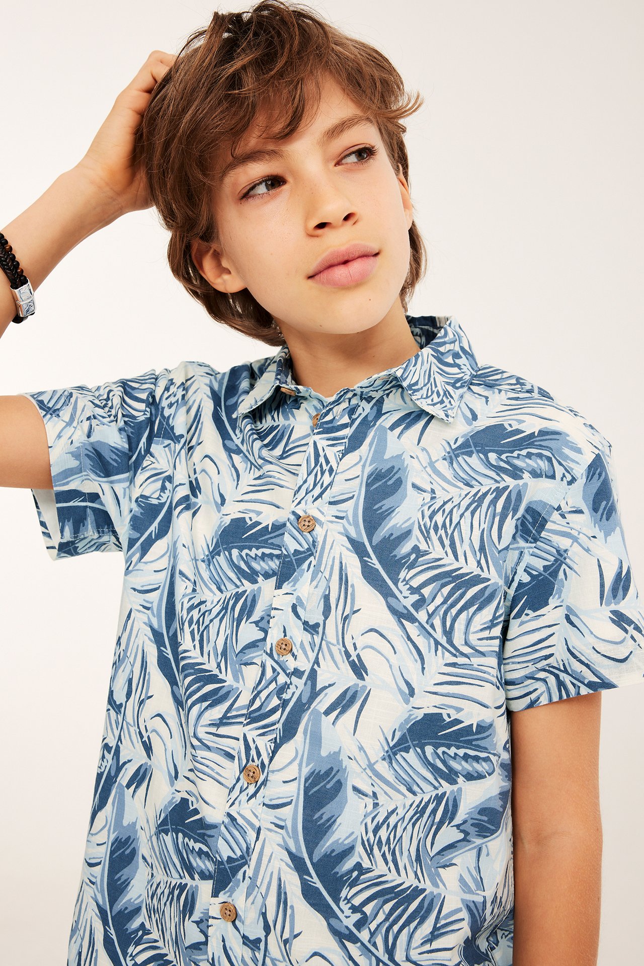 Etunäkymä: Lapsi yllään luonnonvalkoinen lyhythihainen puuvillapaita sinisellä lehtikuviolla ja musta punottu rannekoru.