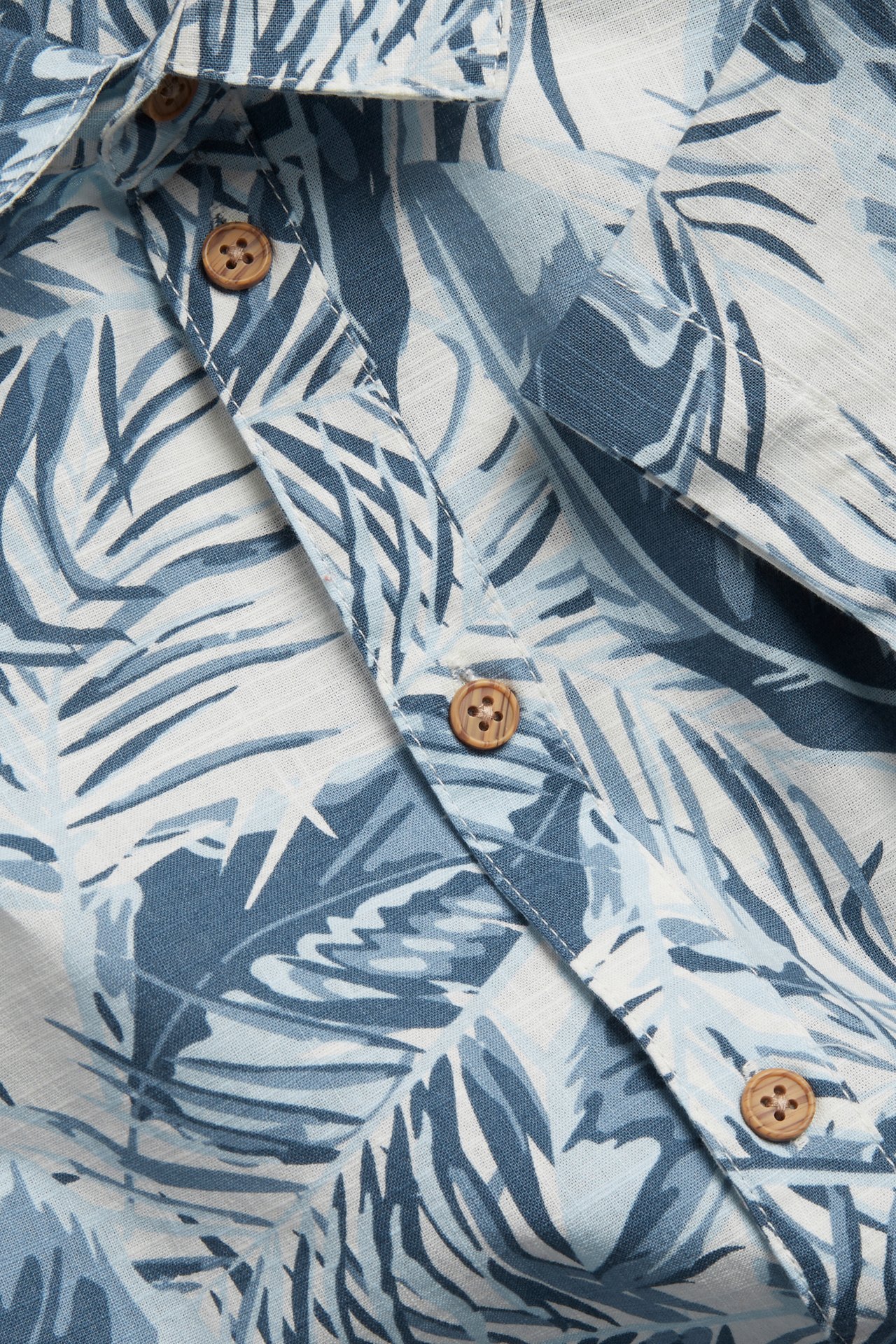 Zbliżenie na koszulę z bawełny ecru z niebieskim wzorem liści palmowych i drewnianymi guzikami.