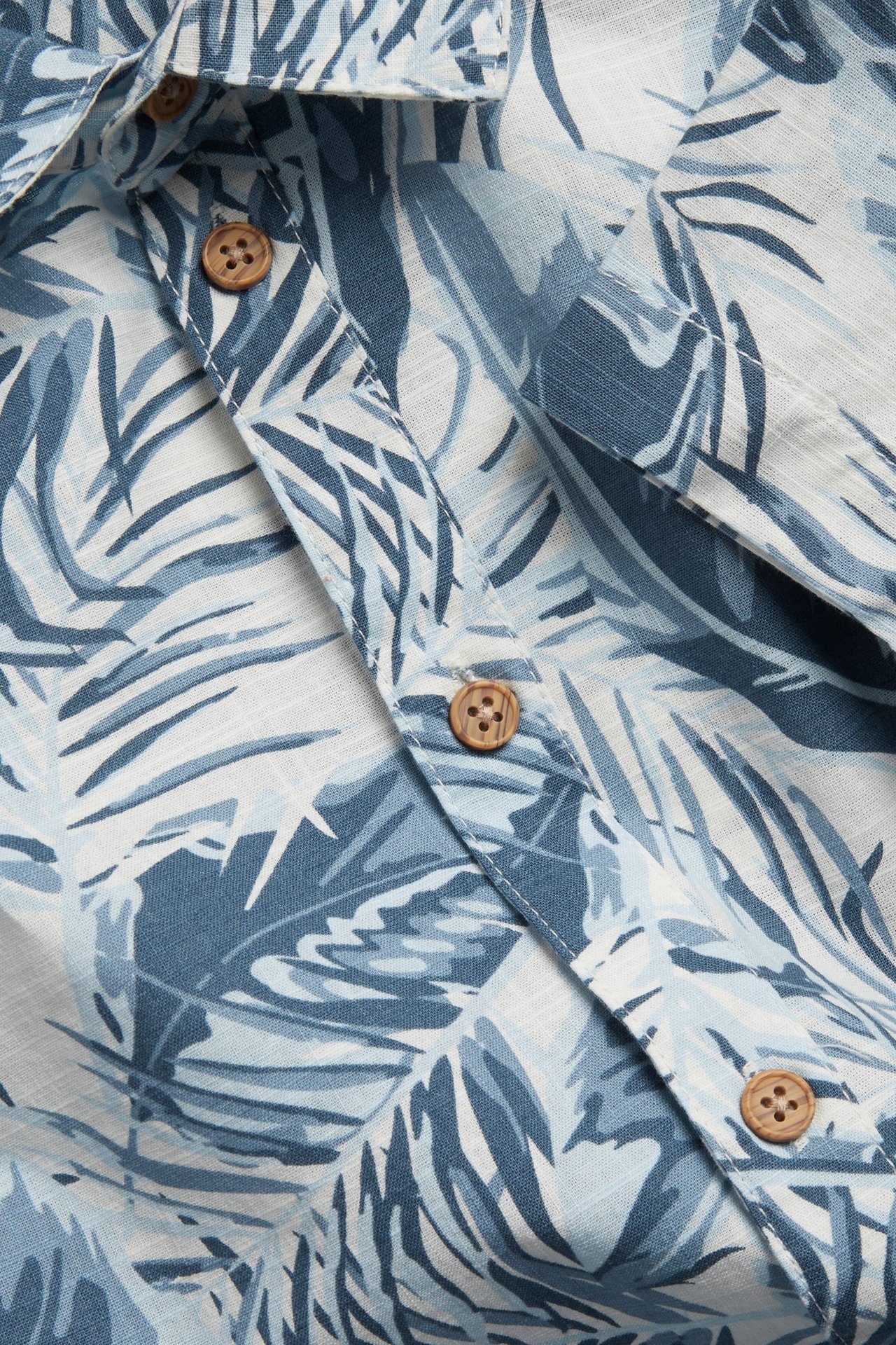 Zbliżenie na koszulę z bawełny ecru z niebieskim wzorem liści palmowych i drewnianymi guzikami.