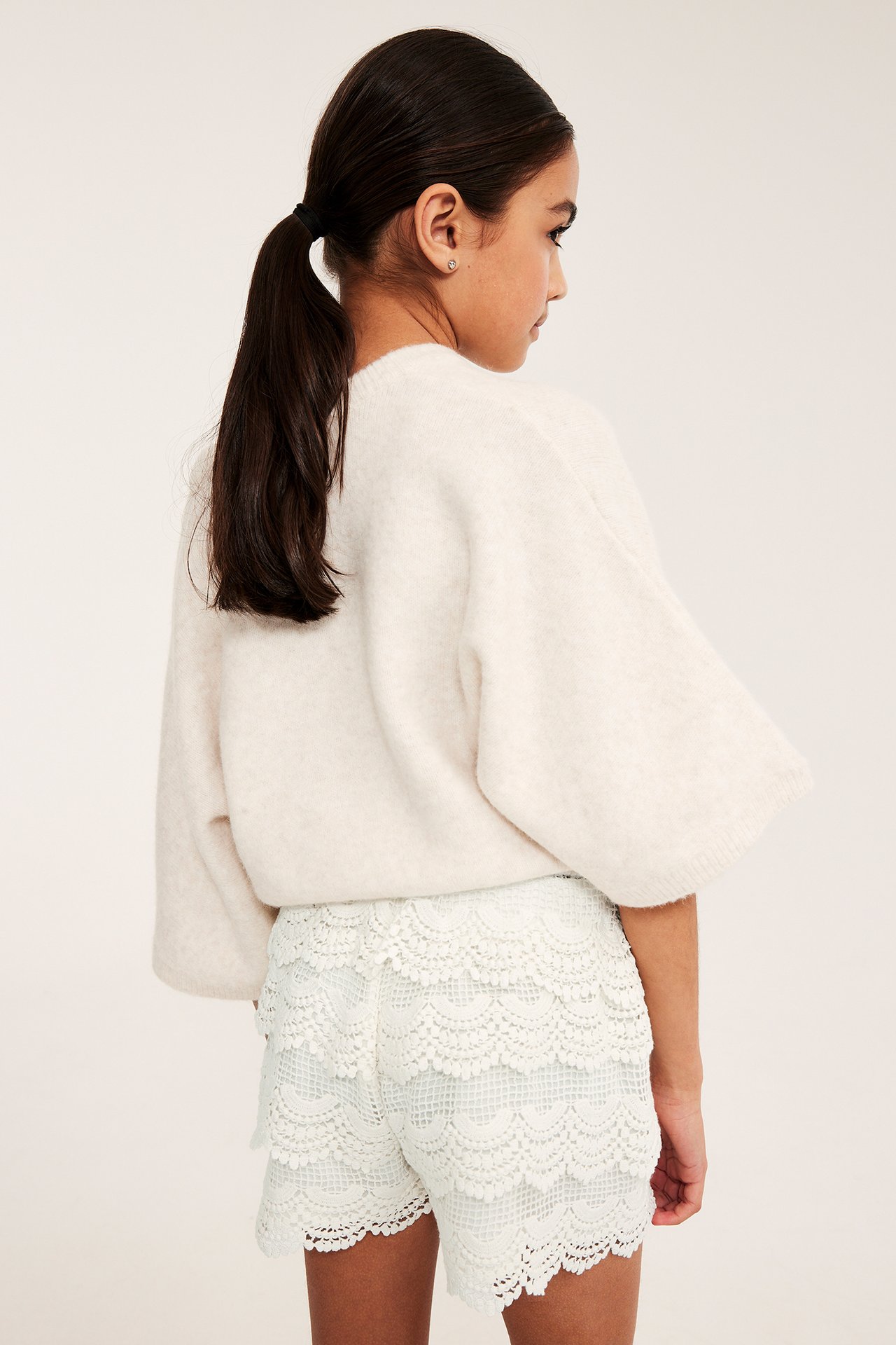 Barns ryggbilde: Offwhite korte blondeshorts med lagdelt bomullsblonder, stylet med en løs offwhite genser.