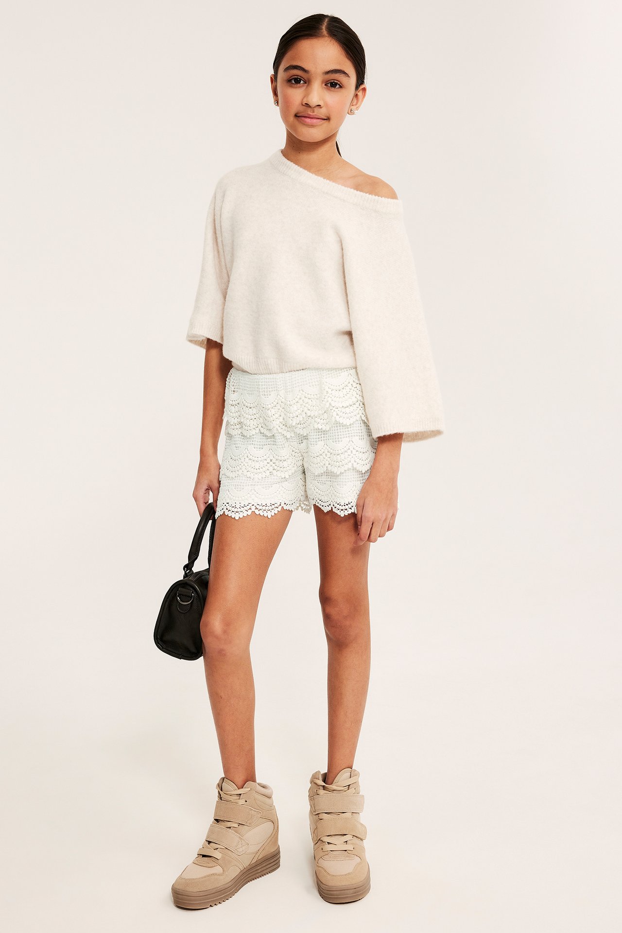 Forfra: Barn i offwhite blondeshorts med strikk i livet, beige strikkegenser, brune sneakers, holder svart veske.