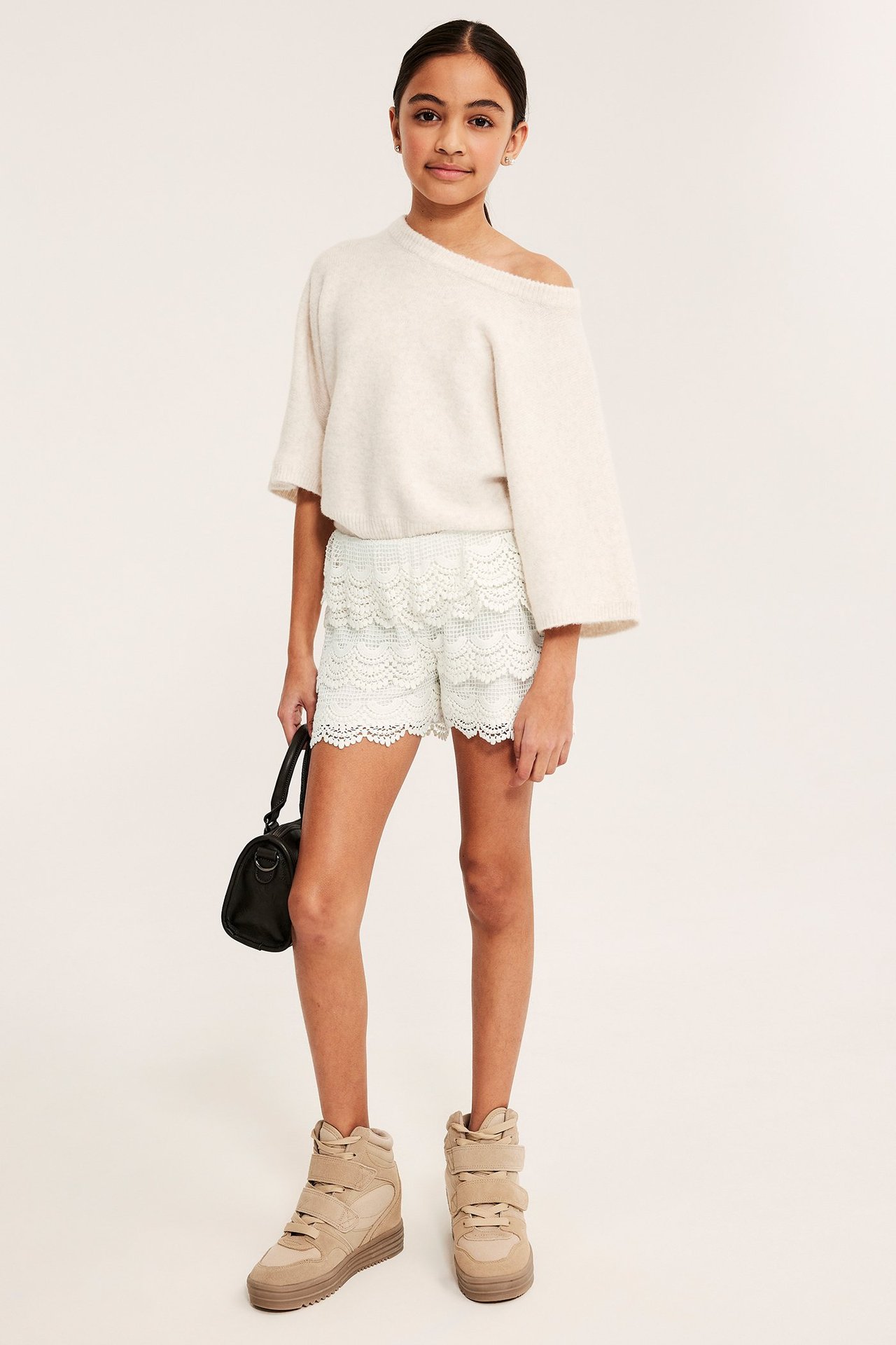Forfra: Barn i offwhite blondeshorts med strikk i livet, beige strikkegenser, brune sneakers, holder svart veske.