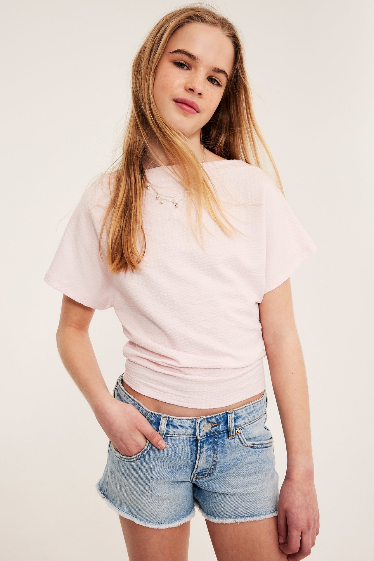 Edestä näkymä lapsesta yllään vaaleat farkkushortsit, joissa on hapsutettu helma, vaaleanpunainen paita ja hopeakaulakoru.