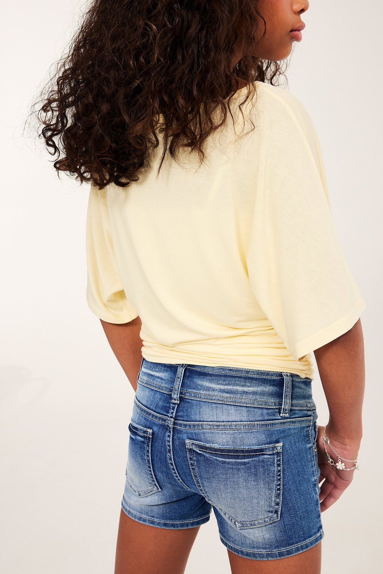 Ryggvy av ett barn i blåa lågmidjade jeansshorts, en gul löst sittande topp och ett silverfärgat blomarmband.