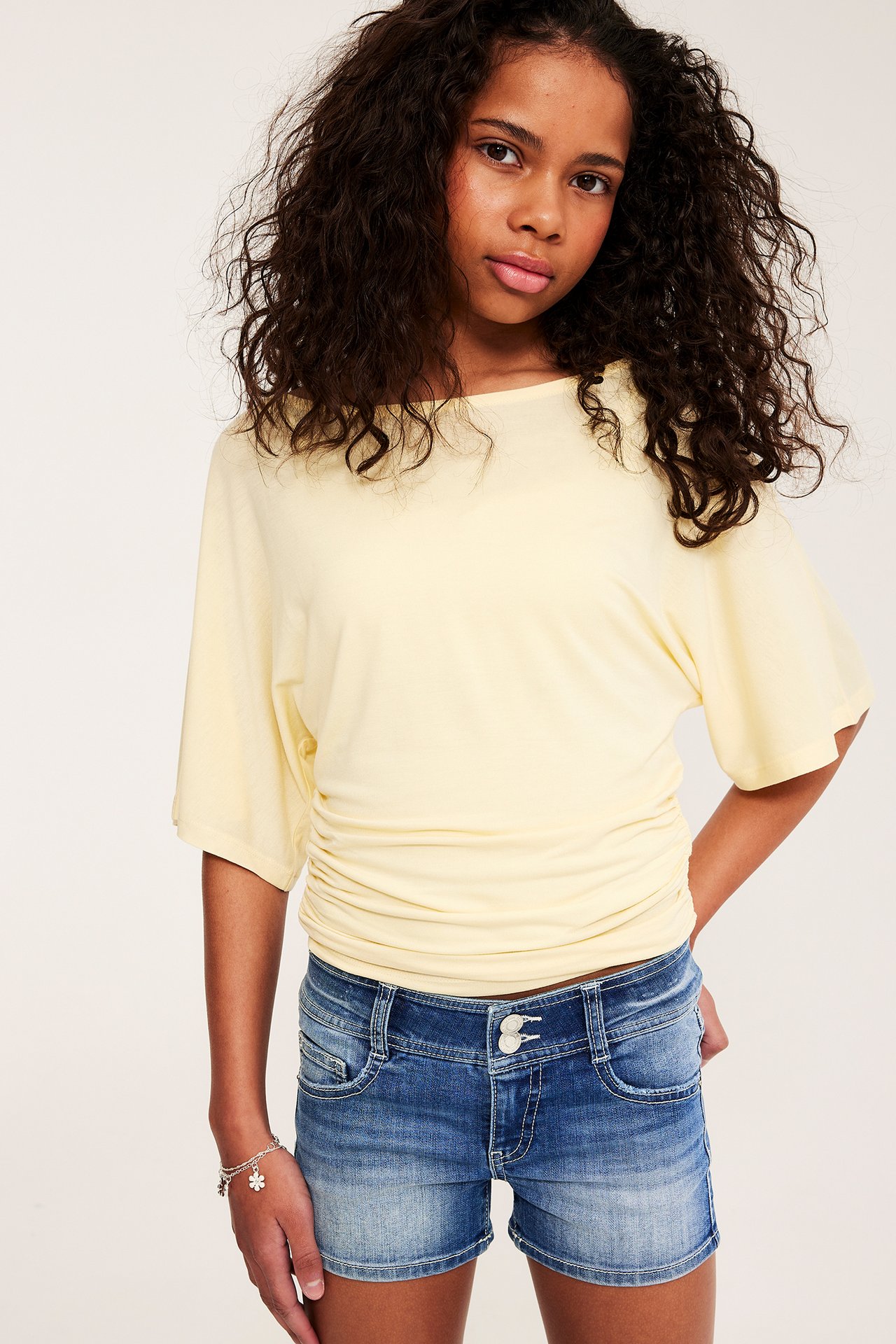 Framifrån: Barn bär blå lågmidjade stretchjeansshorts, en ljusgul topp och silverfärgat blommigt armband.