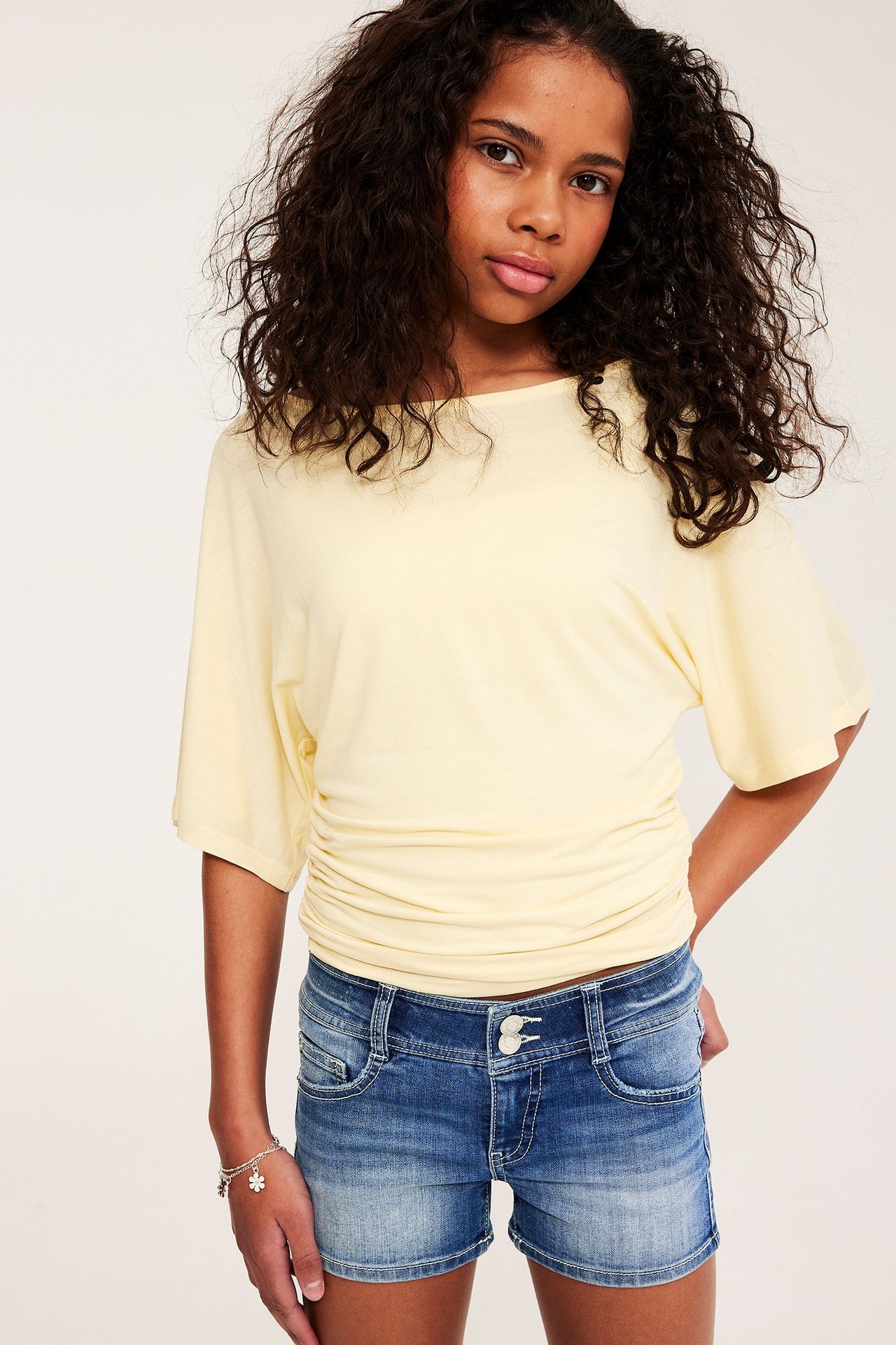 Edestä: Lapsi sinisissä stretch-farkkushortseissa, vaaleankeltaisessa topissa ja hopeisessa rannekorussa.