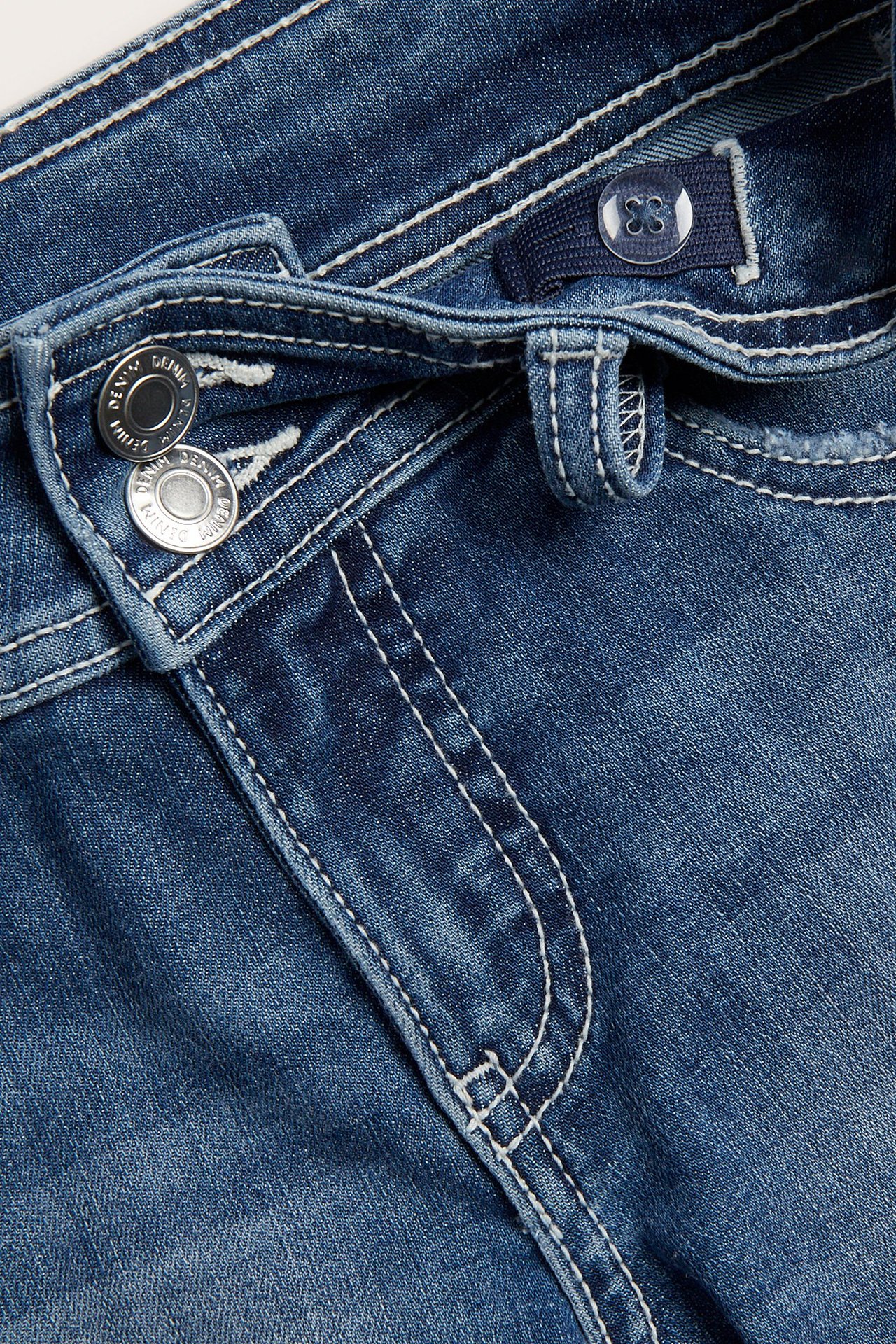 Lähikuva sinisistä matalavyötäröisistä stretchfarkkushortseista, joissa on kaksi hopeanväristä metallinappia ja valkoiset tikkaukset.