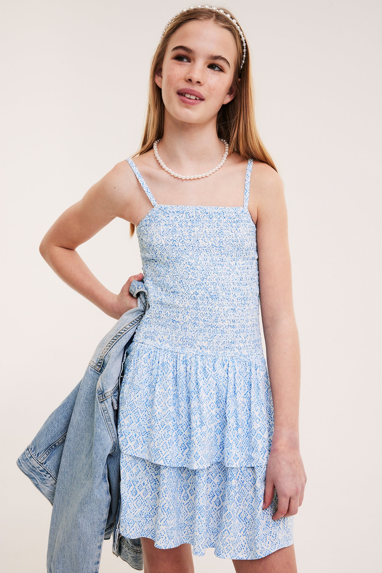 Framsida: Barn i blåmönstrad smockklänning, pärlhalsband, diadem, håller jeansjacka.