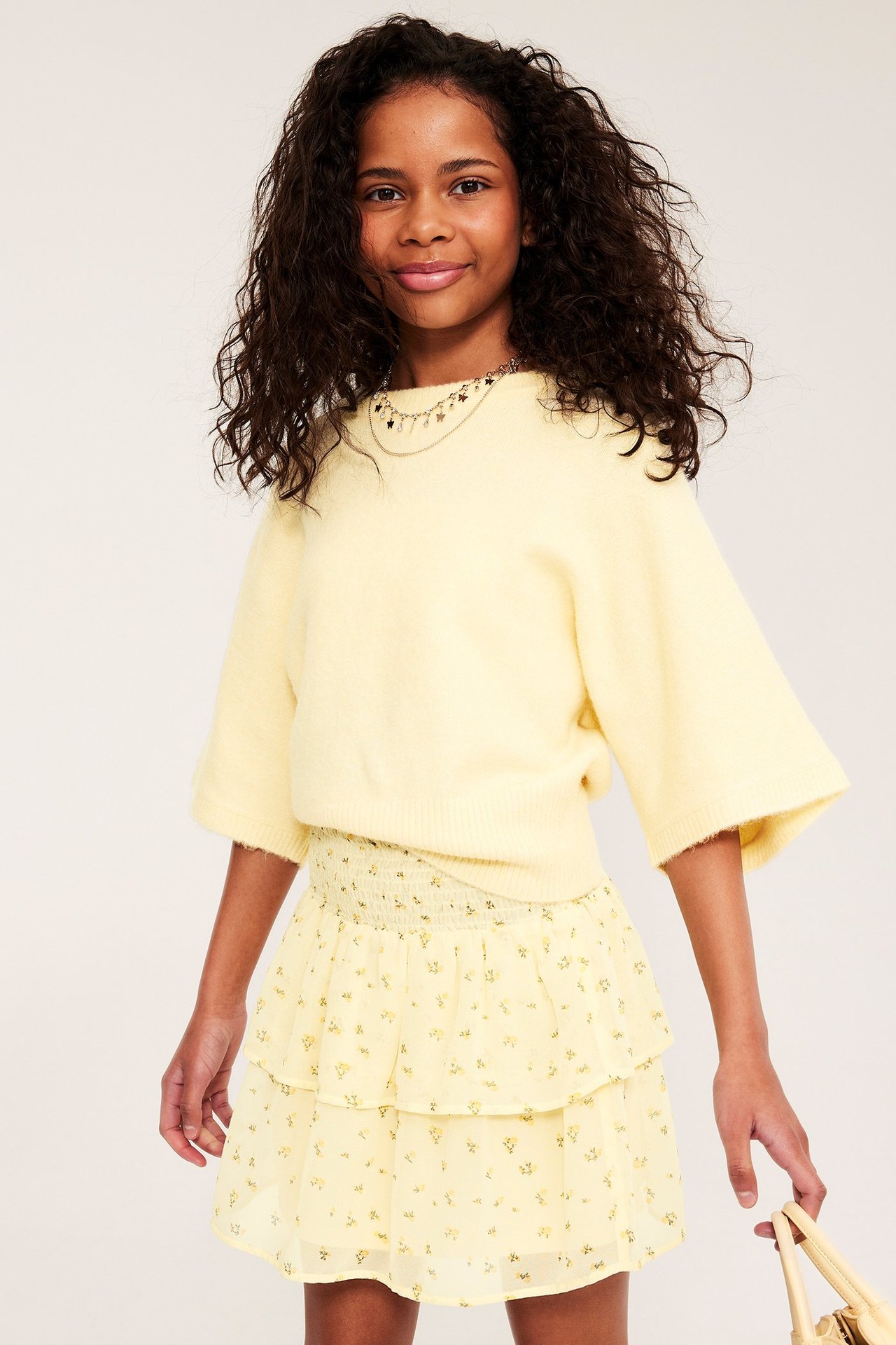 Edestä, lapsi pukeutuneena vaaleankeltaiseen kuvioituun röyhelöhameeseen, jossa smokkirypytys, keltaiseen neuleeseen ja kaulakoruun.