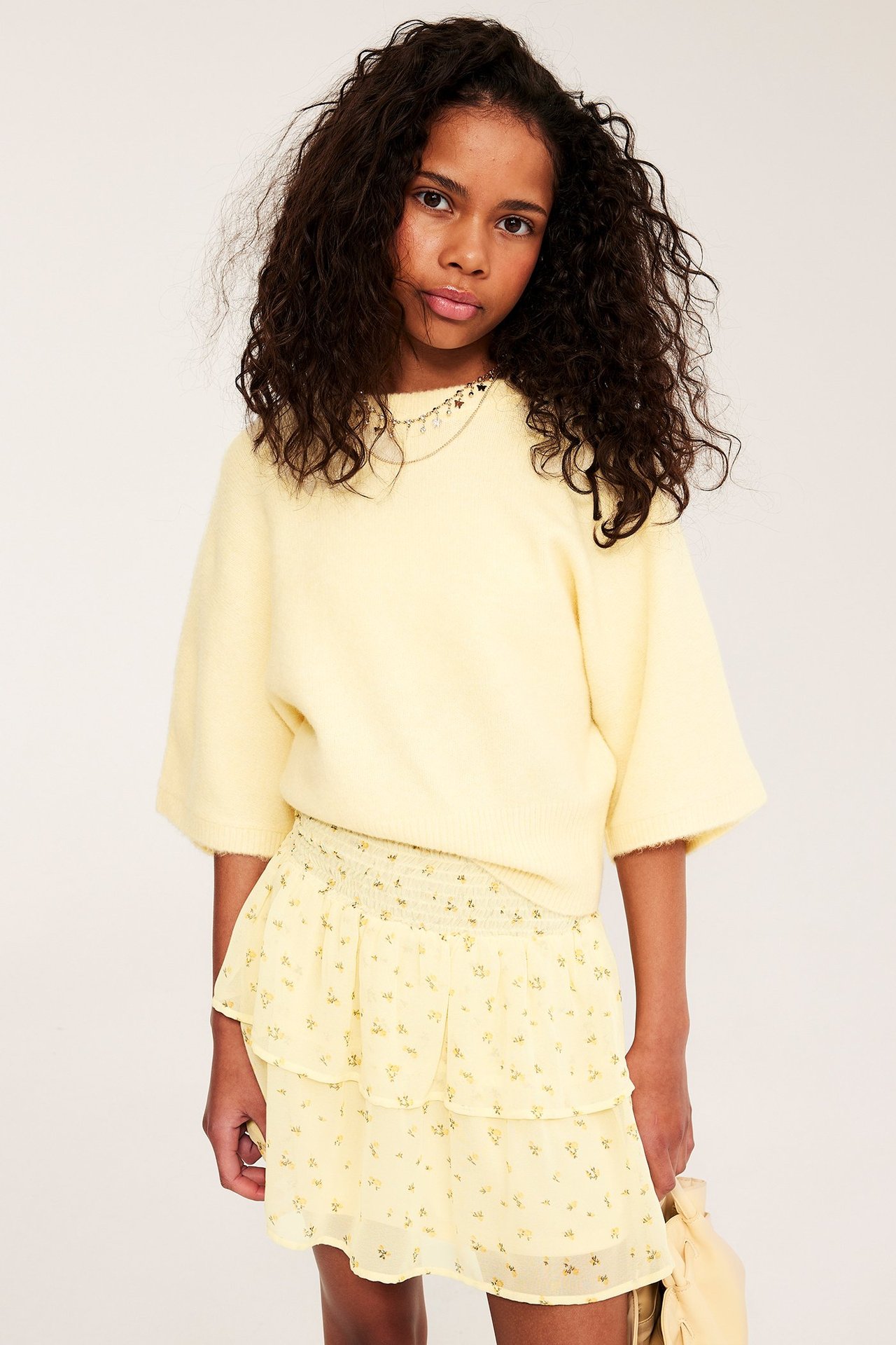 Framifrån: ett barn i ljusgul mönstrad volangkjol med smock och tröja, håller en matchande väska.