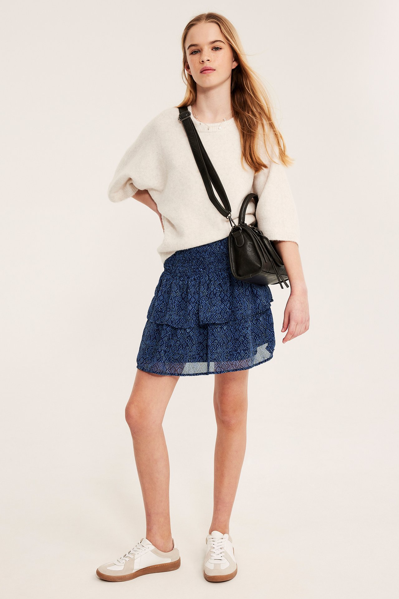 Frontvy av ett barn i mörkblå mönstrad volangkjol med smock och offwhite tröja, med en svart väska.