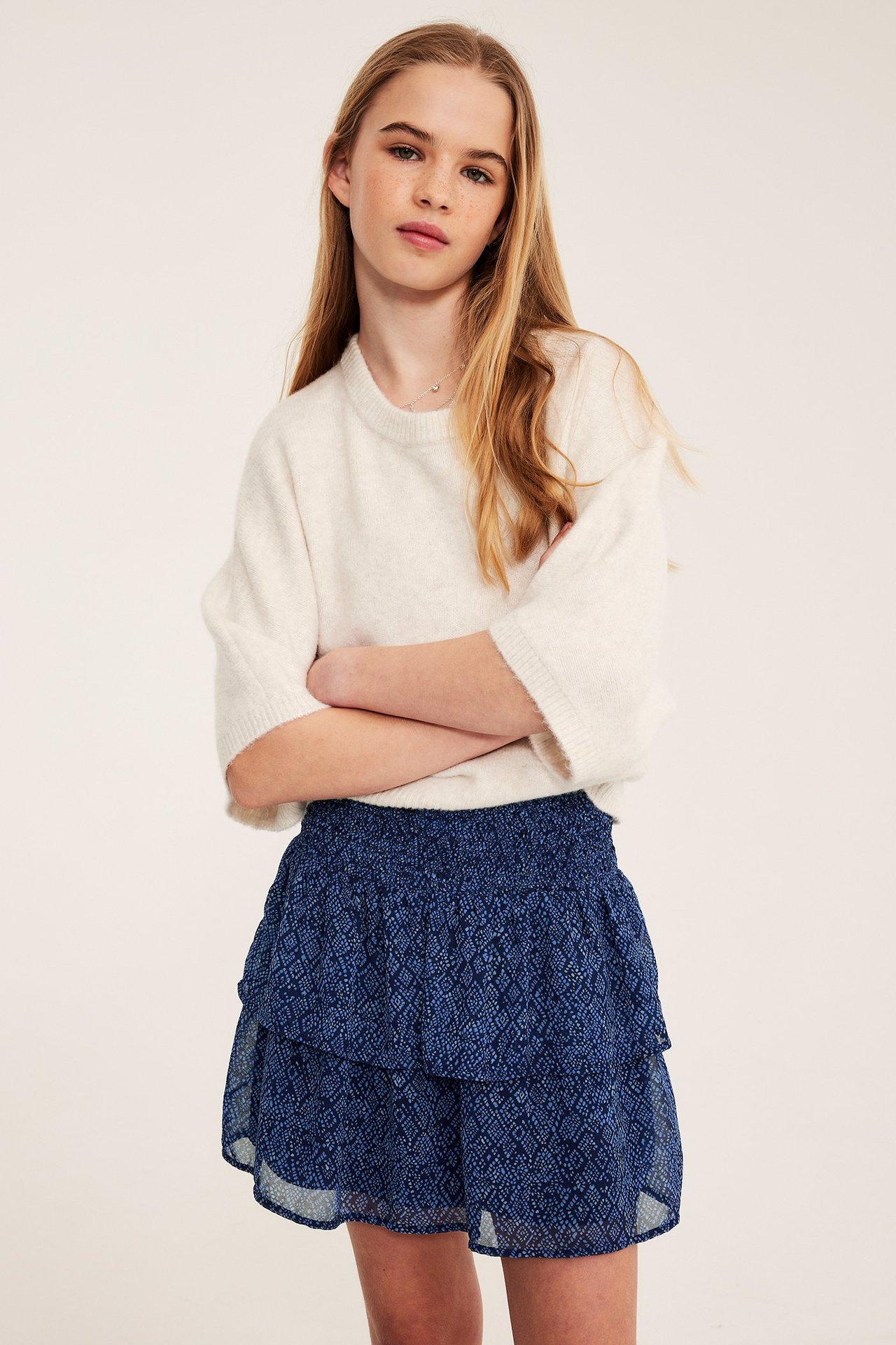 Framifrån: Ett barn bär en mörkblå mönstrad volangkjol med smock och en ljusbeige stickad tröja.