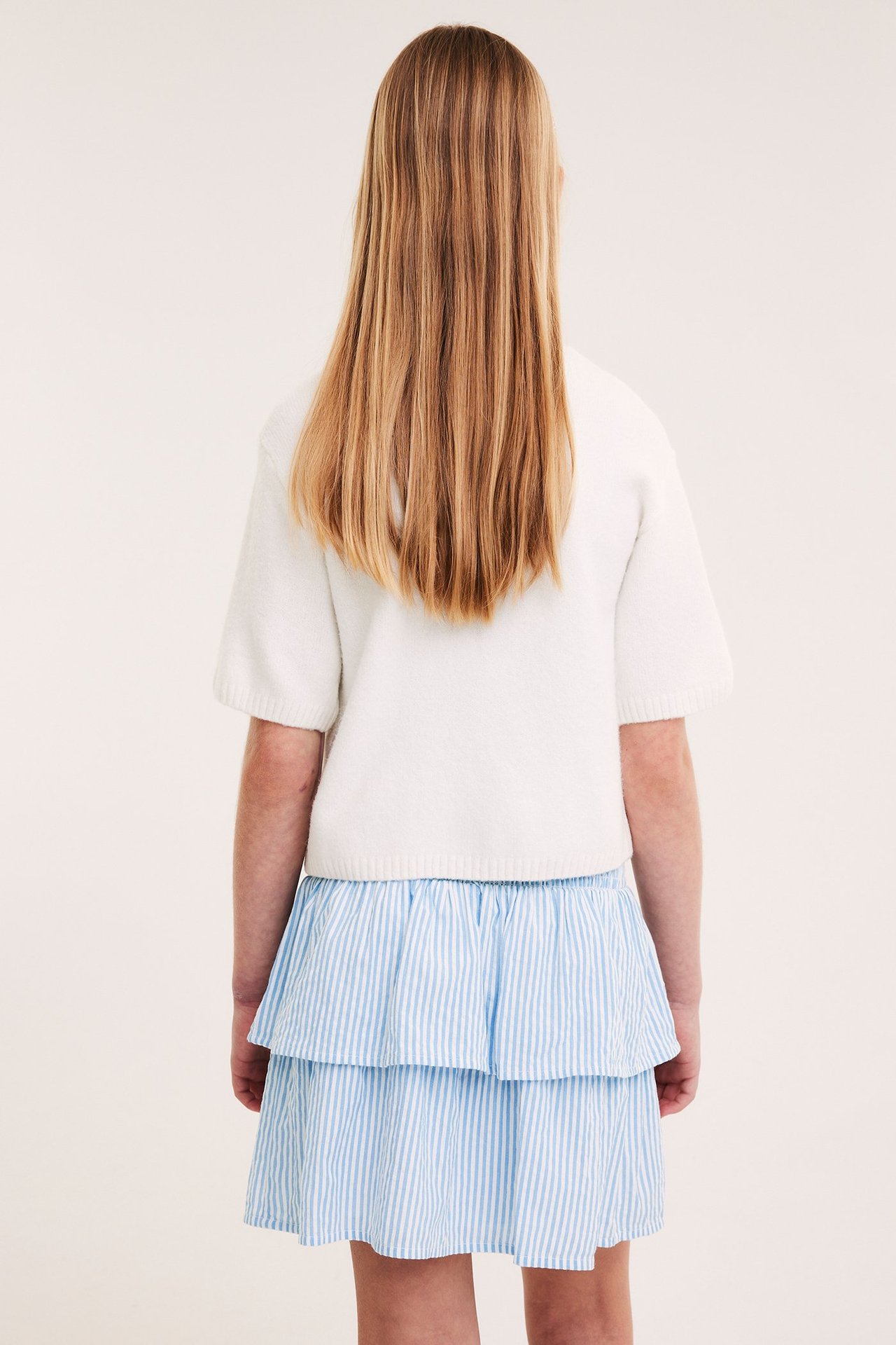Lapsi takaa päin: vaaleansininen ja valkoinen raidallinen röyhelöhame ja valkoinen lyhythihainen toppi.