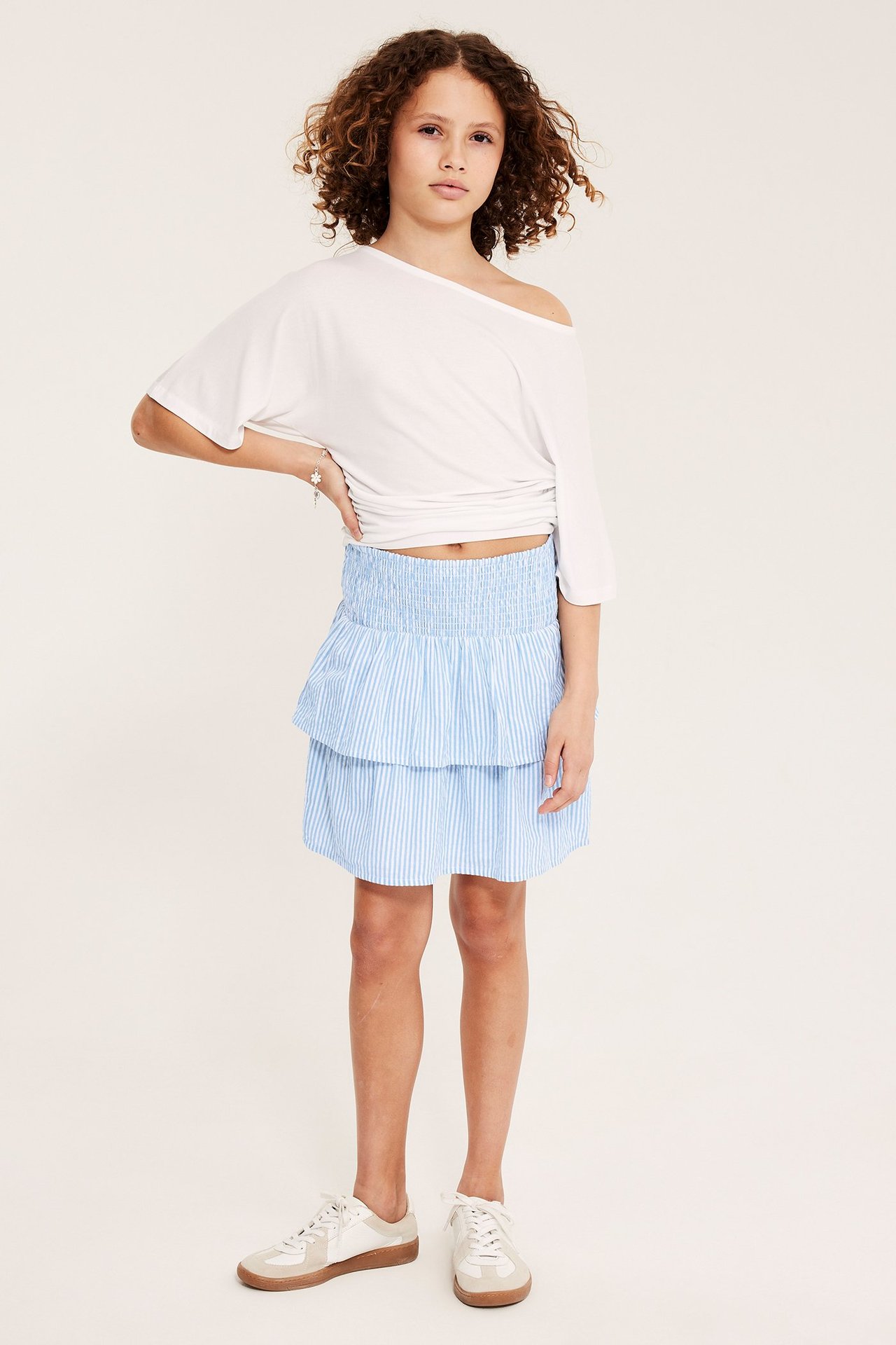 Barn i blåvitrandig volangkjol, vit off-shoulder topp och vita sneakers.