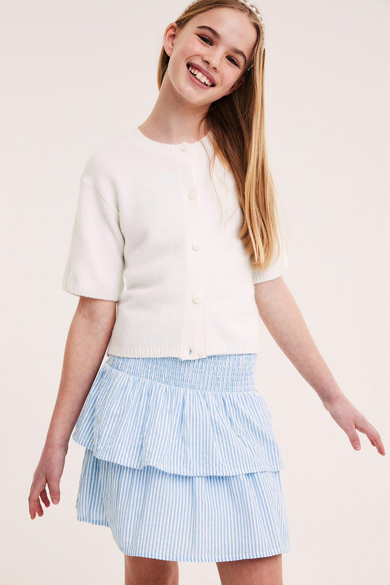 Frontutsikt av et smilende barn i blå- og hvitstripet volangkjole, en hvit kortermet cardigan og et perlehårbånd.