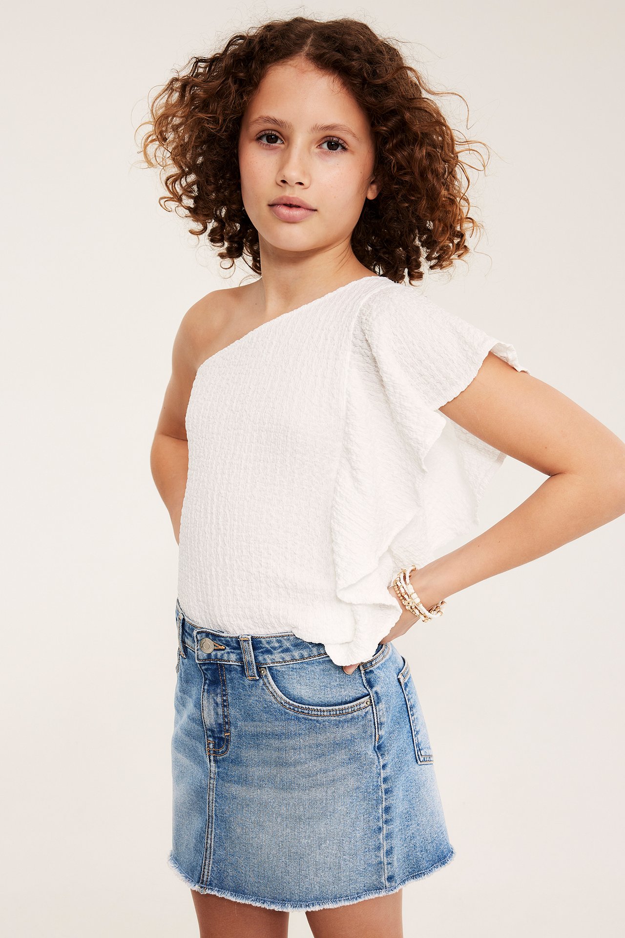 Frontvy av ett barn i ljusblå jeanskjol, vit enaxlad topp och pärlarmband.