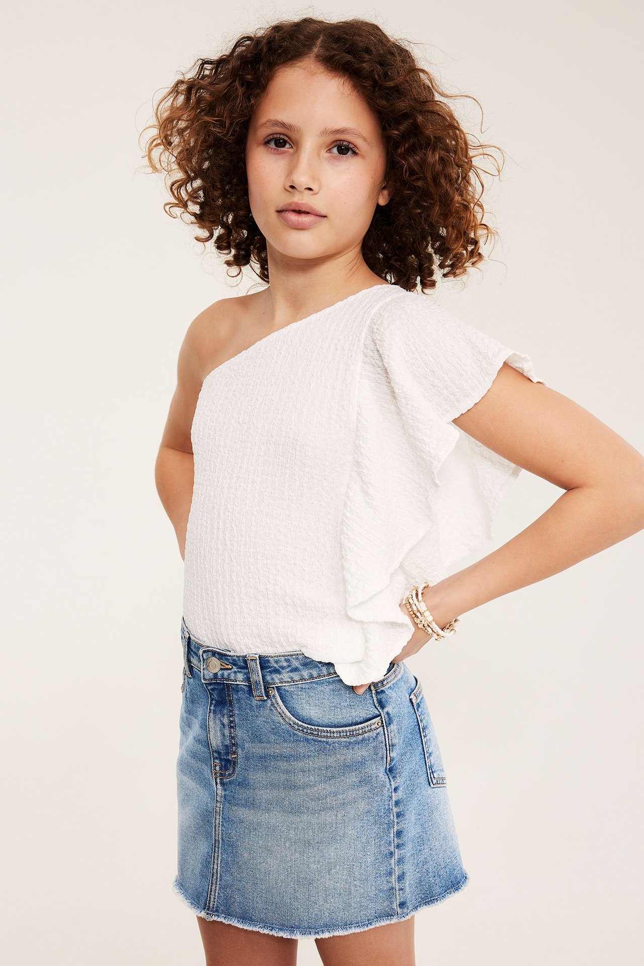 Frontvy av ett barn i ljusblå jeanskjol, vit enaxlad topp och pärlarmband.