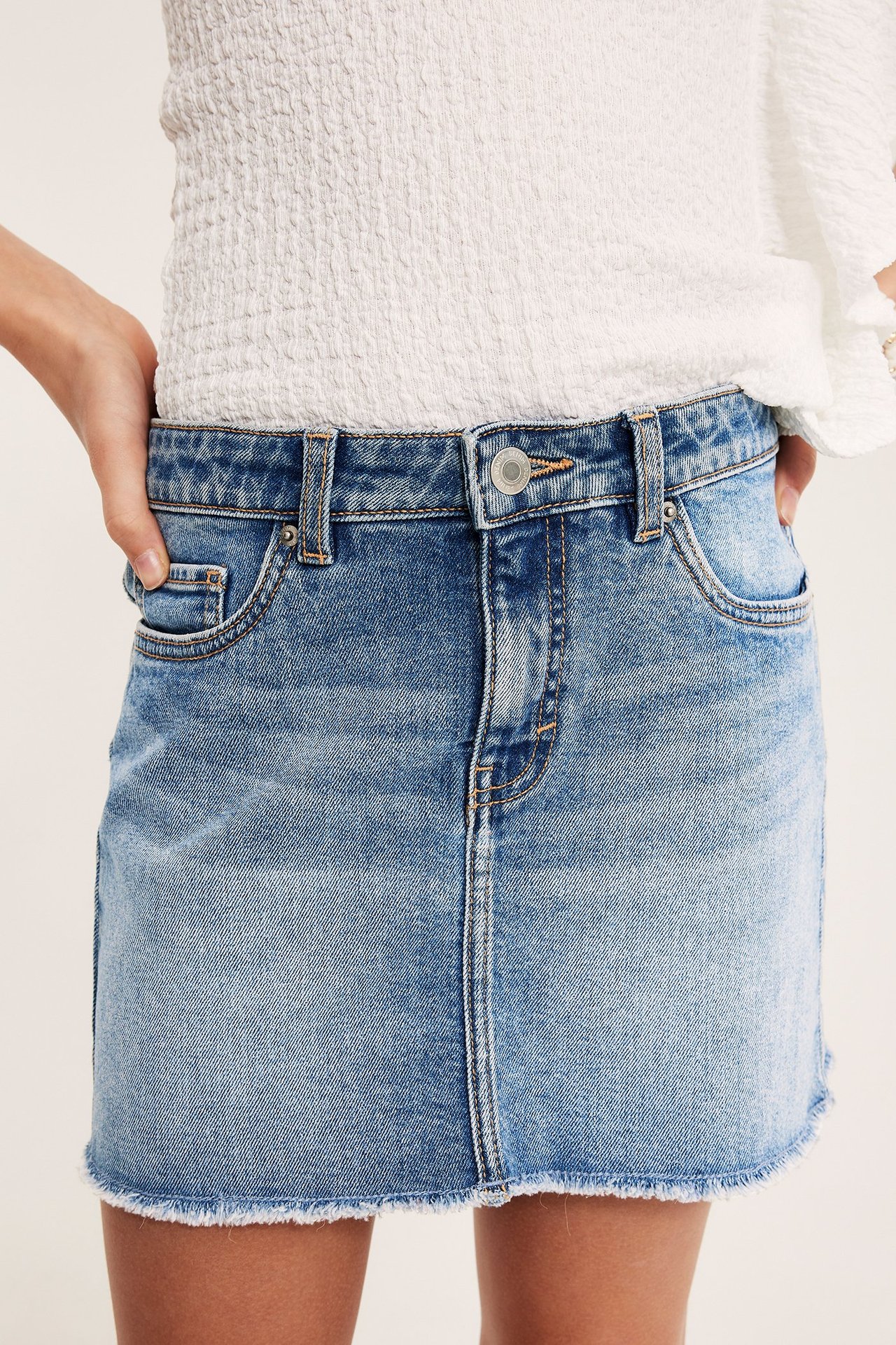 Frontvisning av et barn i lyseblått lavmidjet dongeriskjørt med frynset kant og hvit smock-topp.