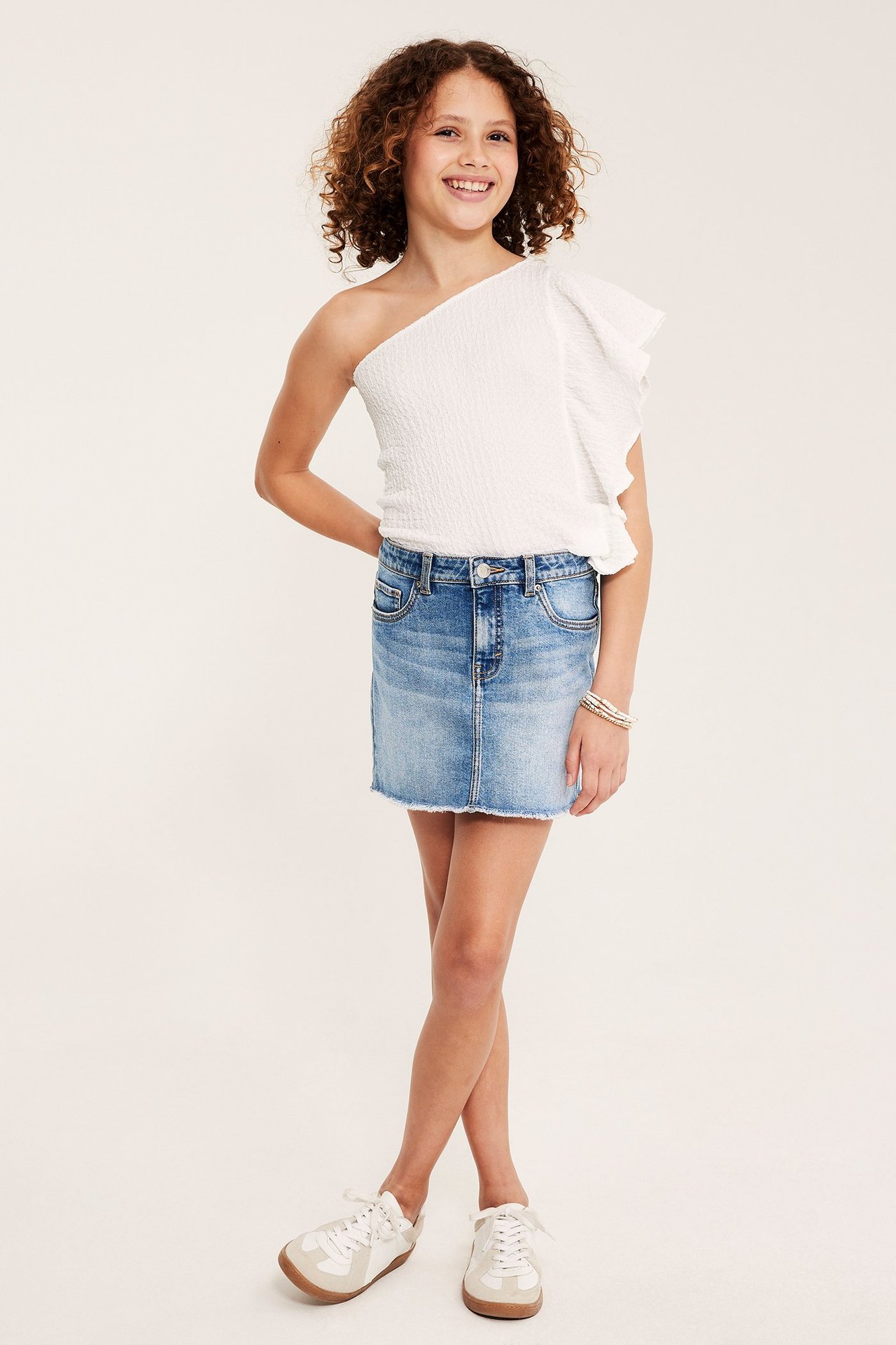 Framifrån: Tjej i ljusblå jeanskjol med låg midja, vit enaxlad topp och vita sneakers.