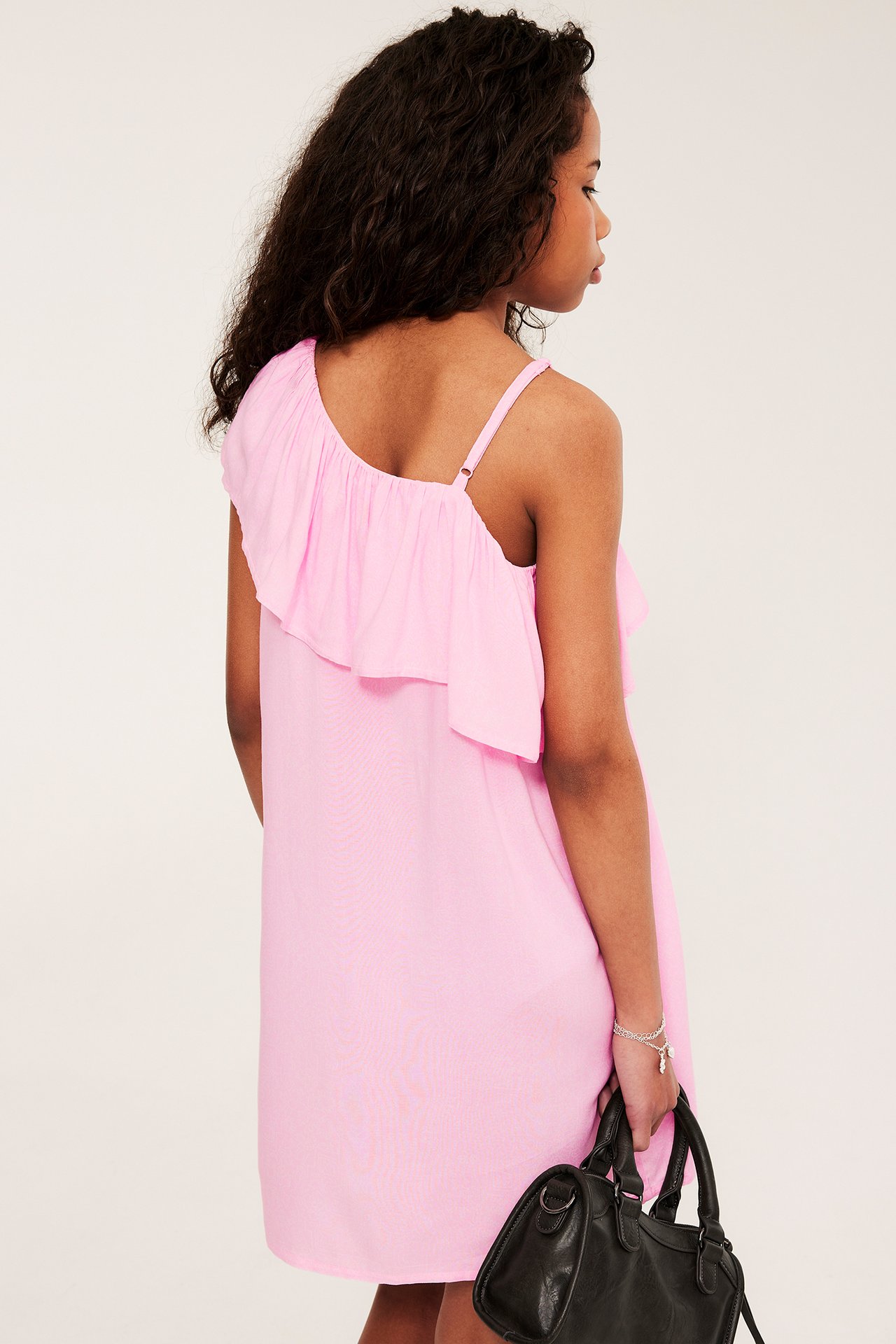 Dziecko z tyłu w różowej sukience na jedno ramię z falbanką, z czarną torebką i srebrną bransoletką.