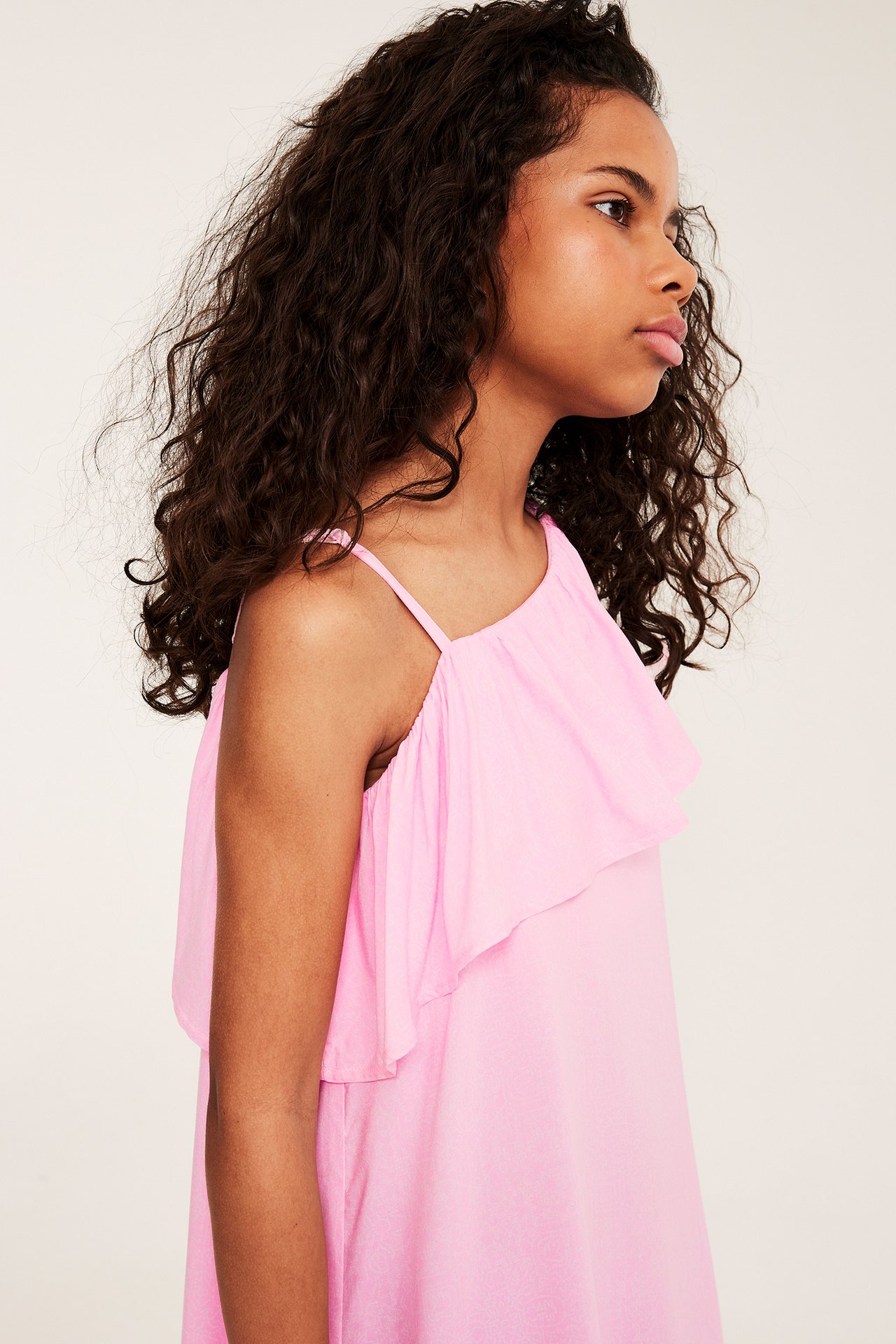 Sidevisning av et barn i en rosa viskose one-shoulder kjole med rysjekant.