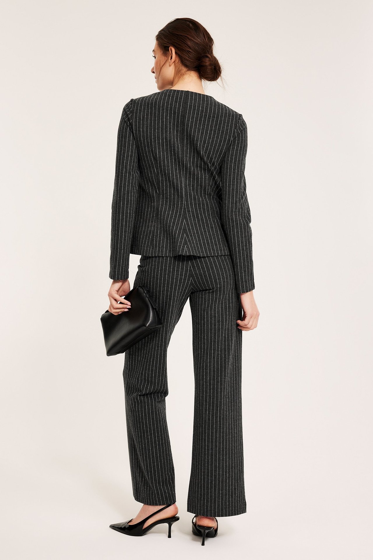 Widok z tyłu: kobieta w ciemnoszarym garniturze w prążki z szerokimi nogawkami, trzyma czarną kopertówkę, nosi czarne czółenka.