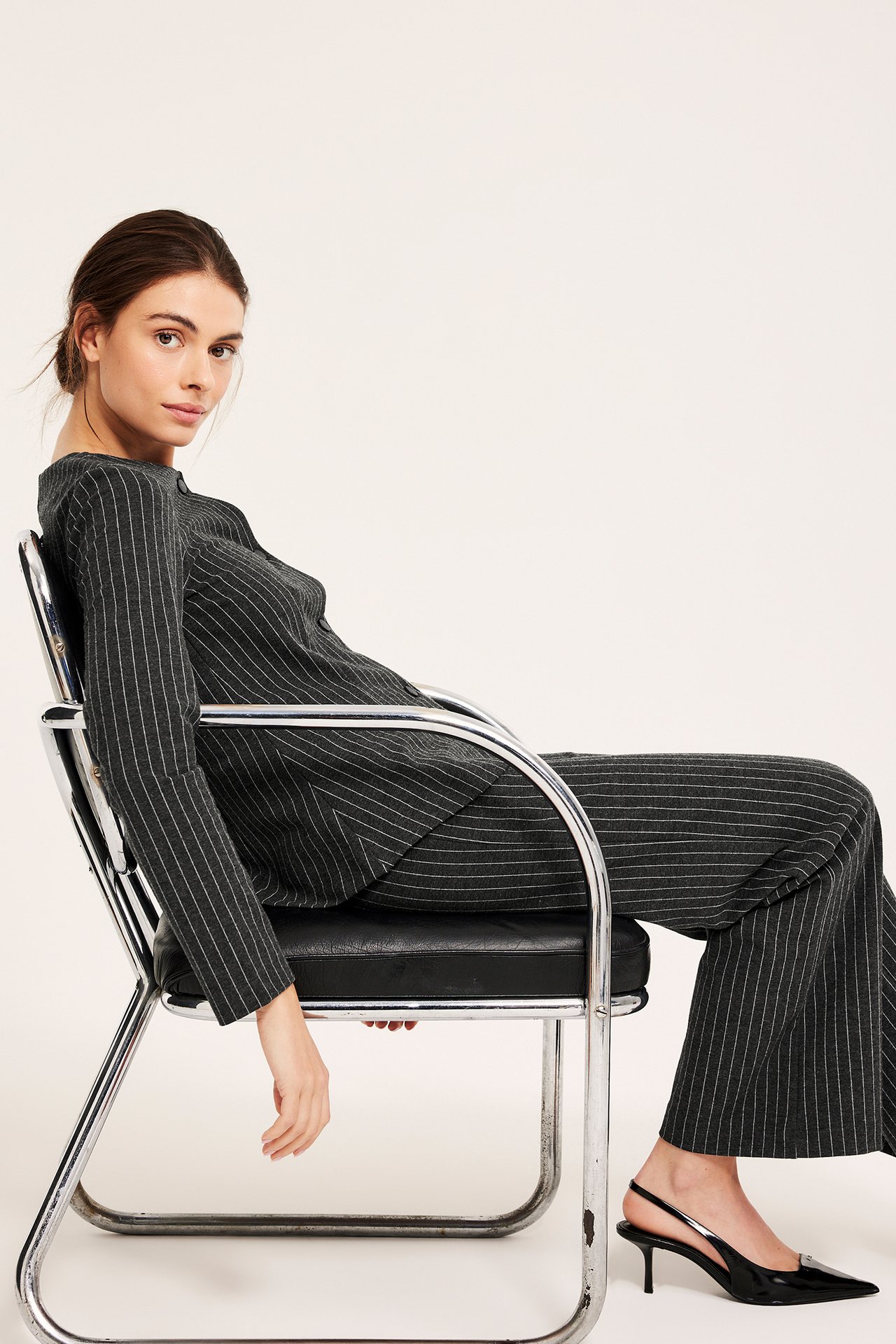 Widok z boku kobiety w ciemnoszarych spodniach w paski, dopasowanym topie z długim rękawem i czarnych szpilkach.