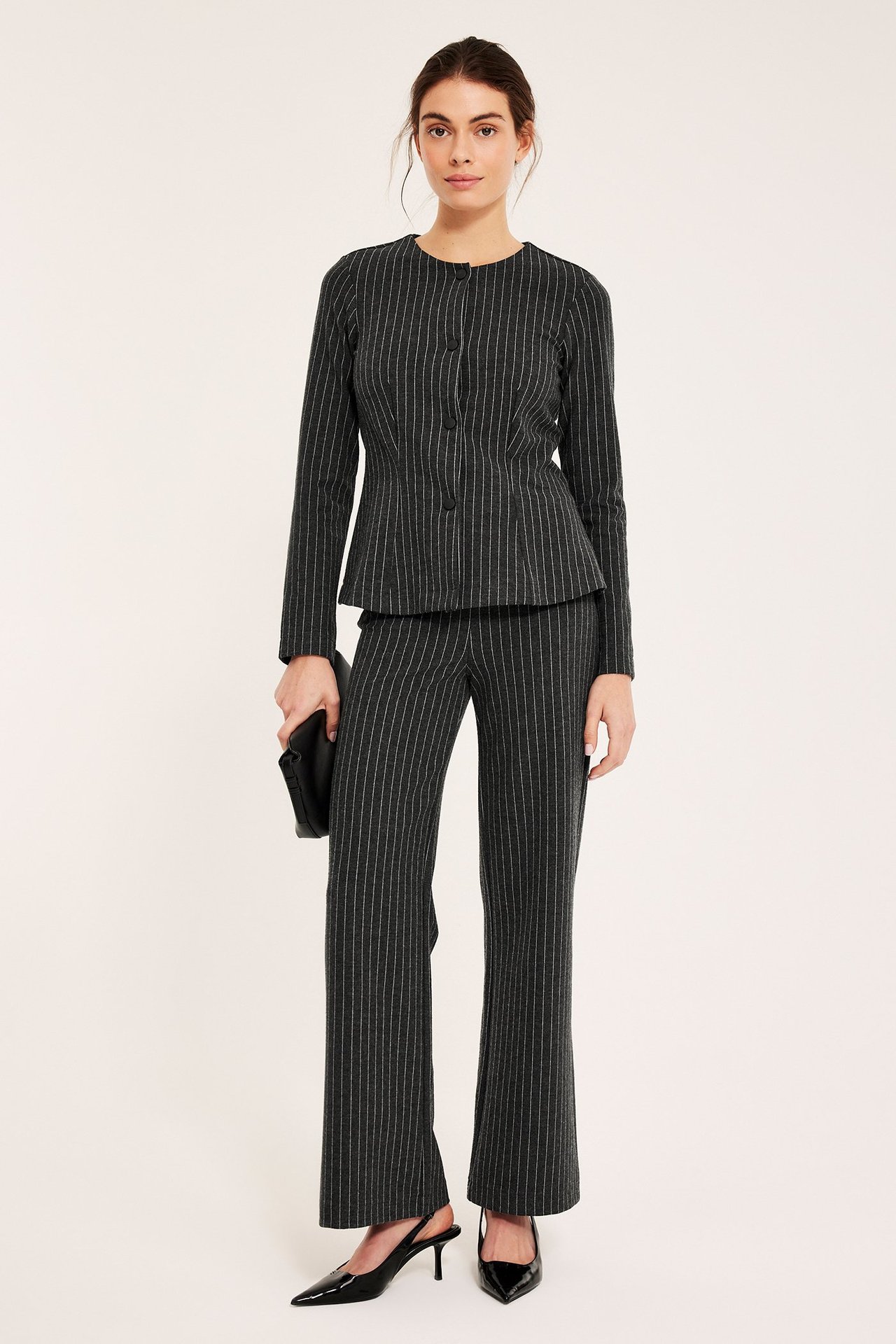 Kvinne i mørkegrå nålestripete vide bukser, matchende jakke, svart clutch og hæler, frontutsikt.
